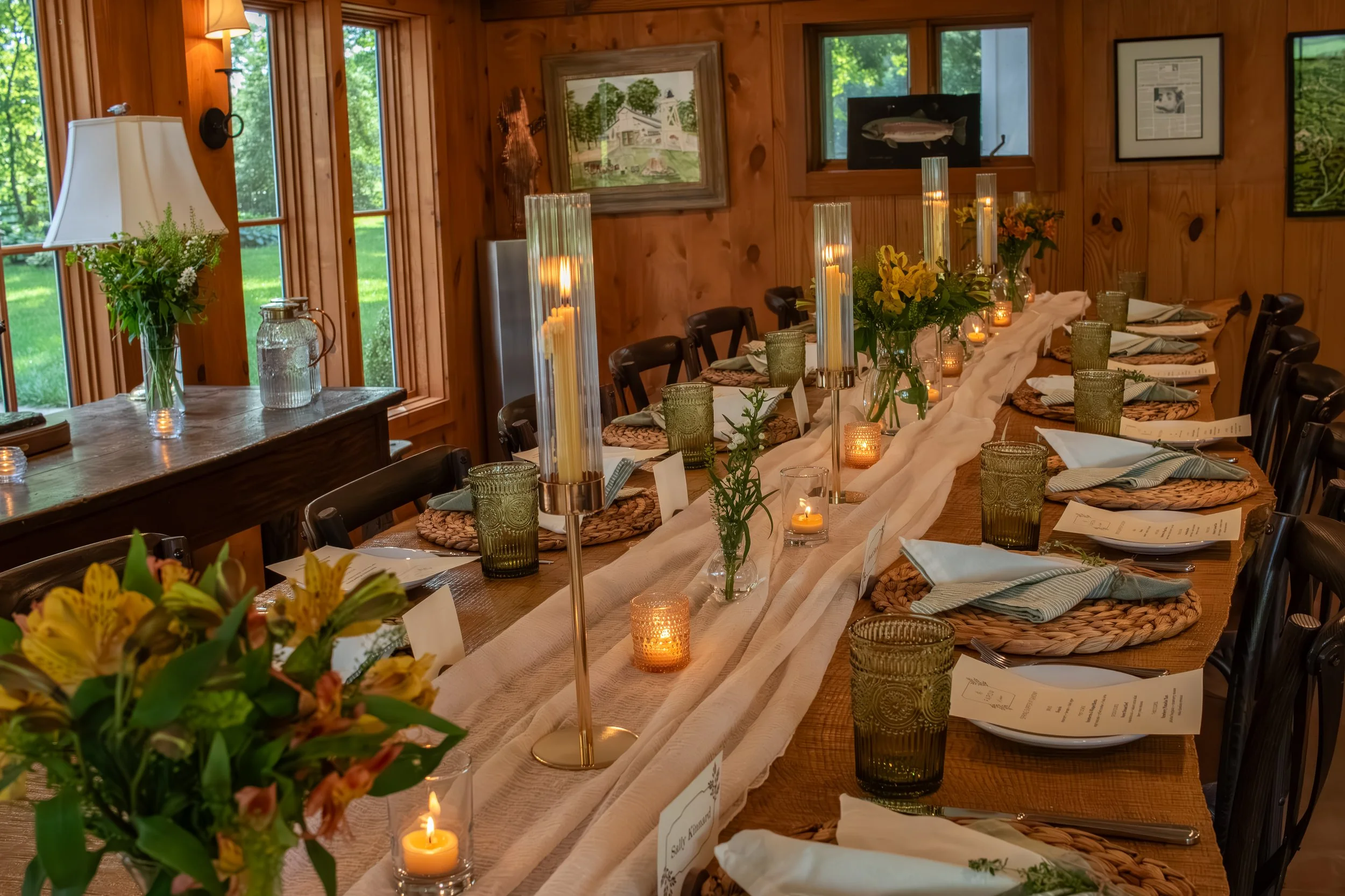 A decorated banquet table with a rustic wooden interior, featuring a floral centerpiece, multiple candles, placed settings with napkins, several glasses, and framed pictures on the wall behind, with windows showing a green outdoor landscape.