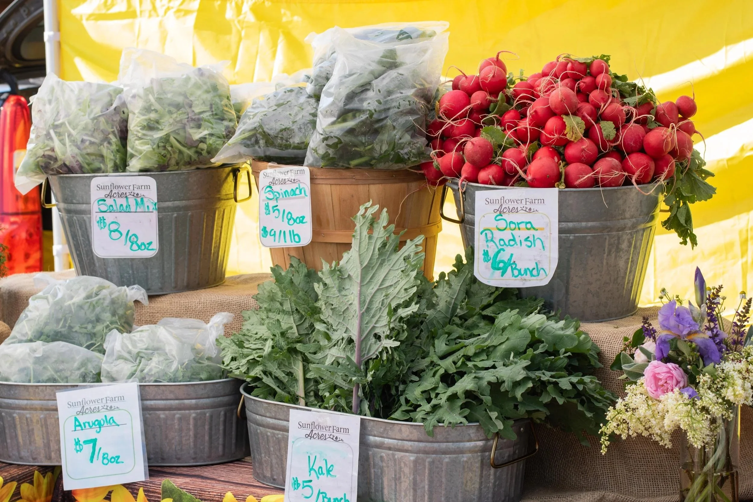 Fresh vegetables and flowers at a market stall. Visible items include radishes, kale, arugula, spinach, and a bouquet of mixed flowers.