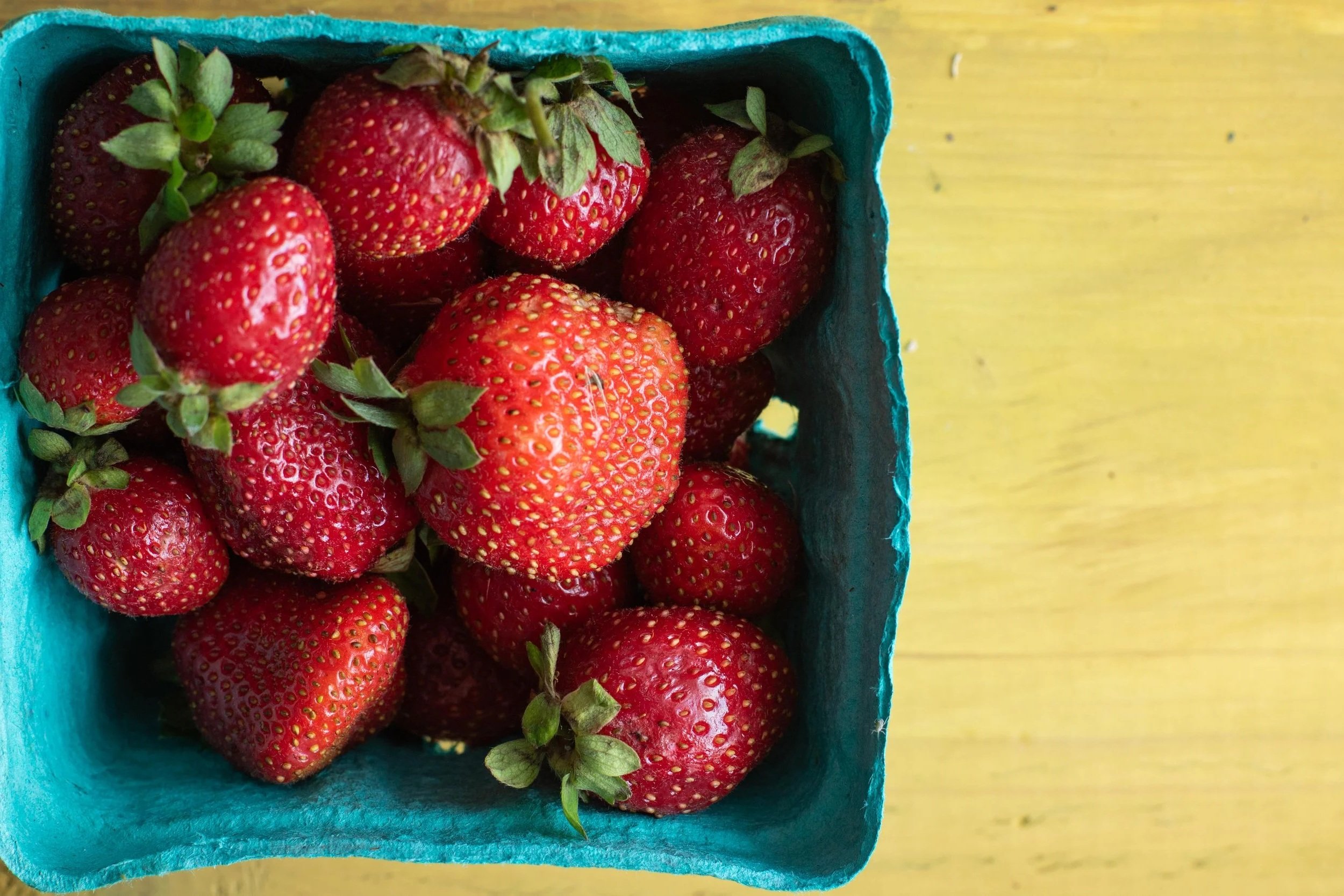 Fresh strawberries in a teal basket on a yellow wooden surface.