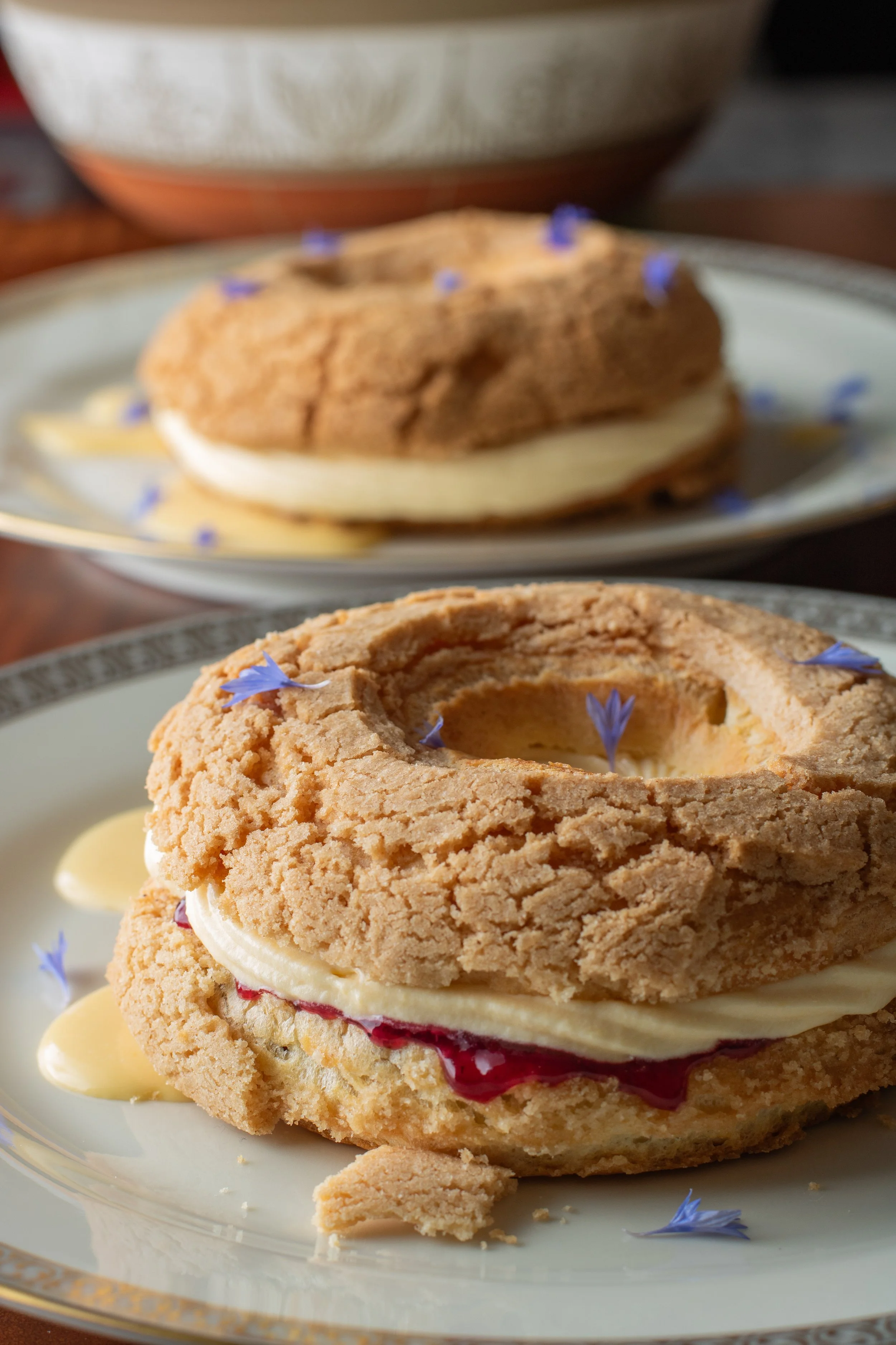 Close-up of a dessert with a crumbly cookie top, layers of cream and red berry jam filling, garnished with small blue flowers, served on a decorative plate.