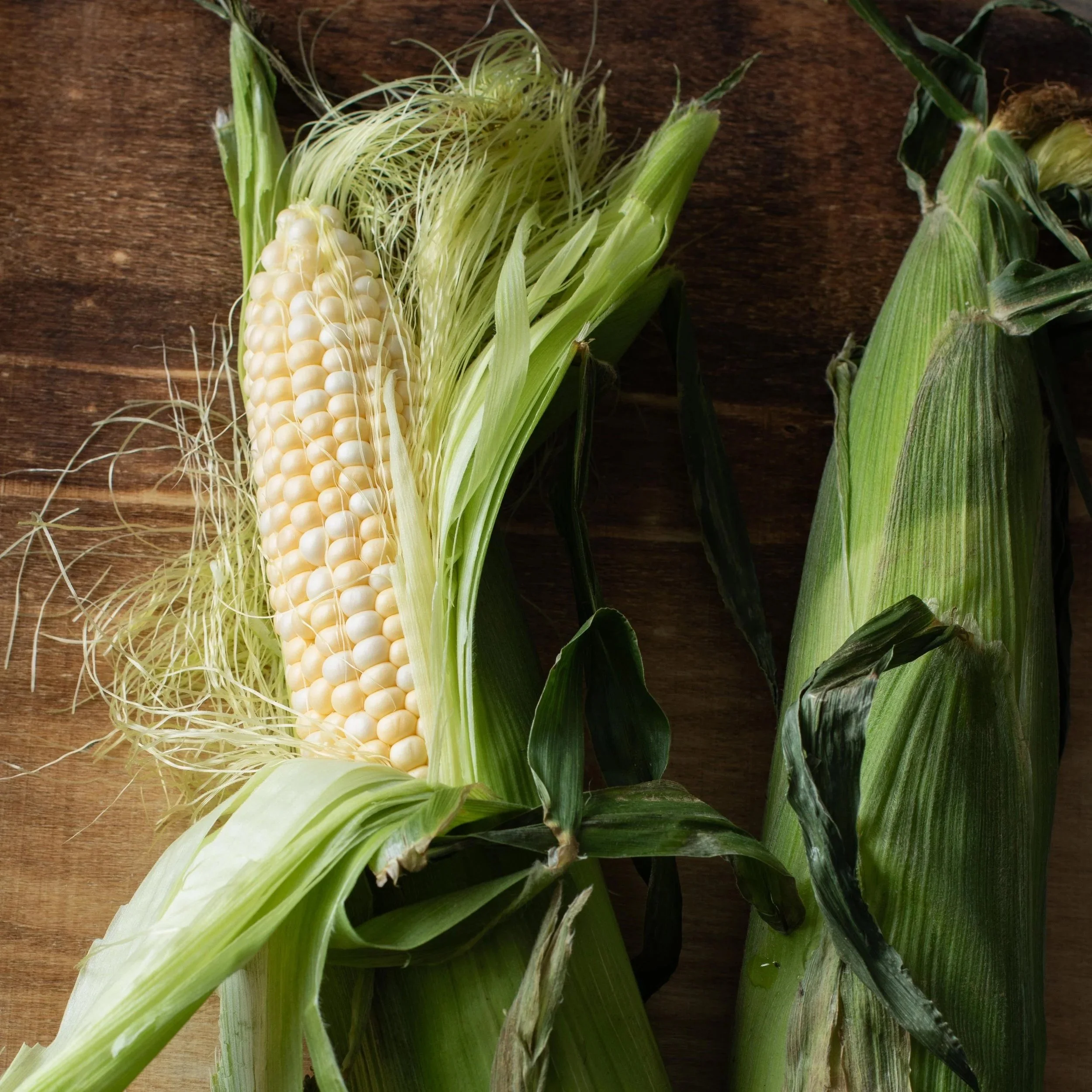 A partially husked ear of white corn on a wooden surface, with green husks and silk visible.