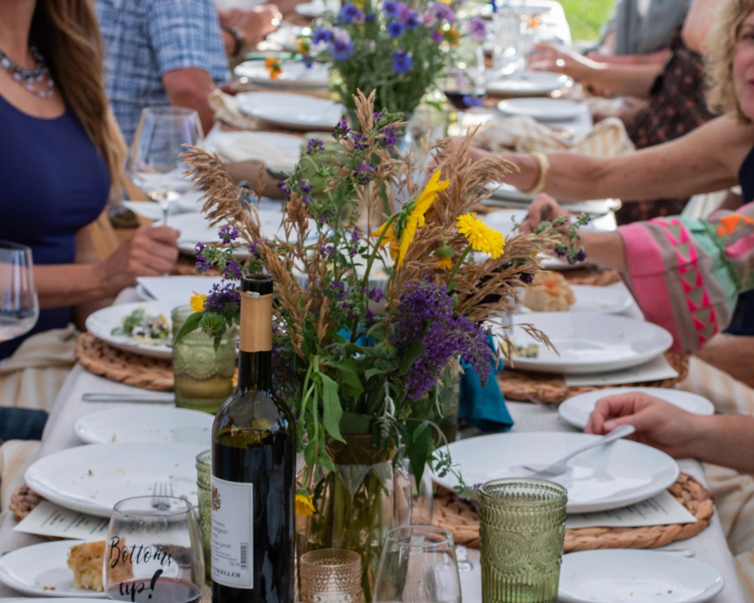 A long dining table set for a gathering with plates, glasses, and a centerpiece of wildflowers, with people partially visible in the background.