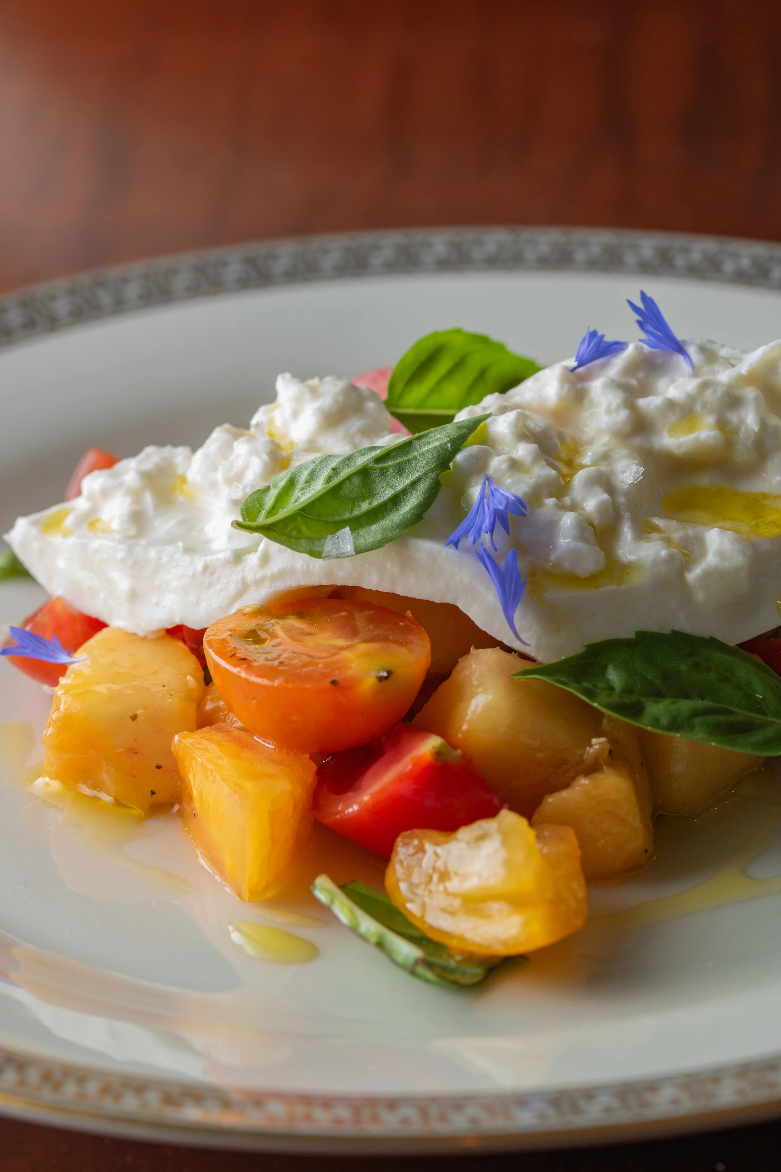 A bowl of fresh fruit salad topped with whipped cream, garnished with basil leaves and edible blue flowers, with a slice of tomato and other diced fruits visible underneath.