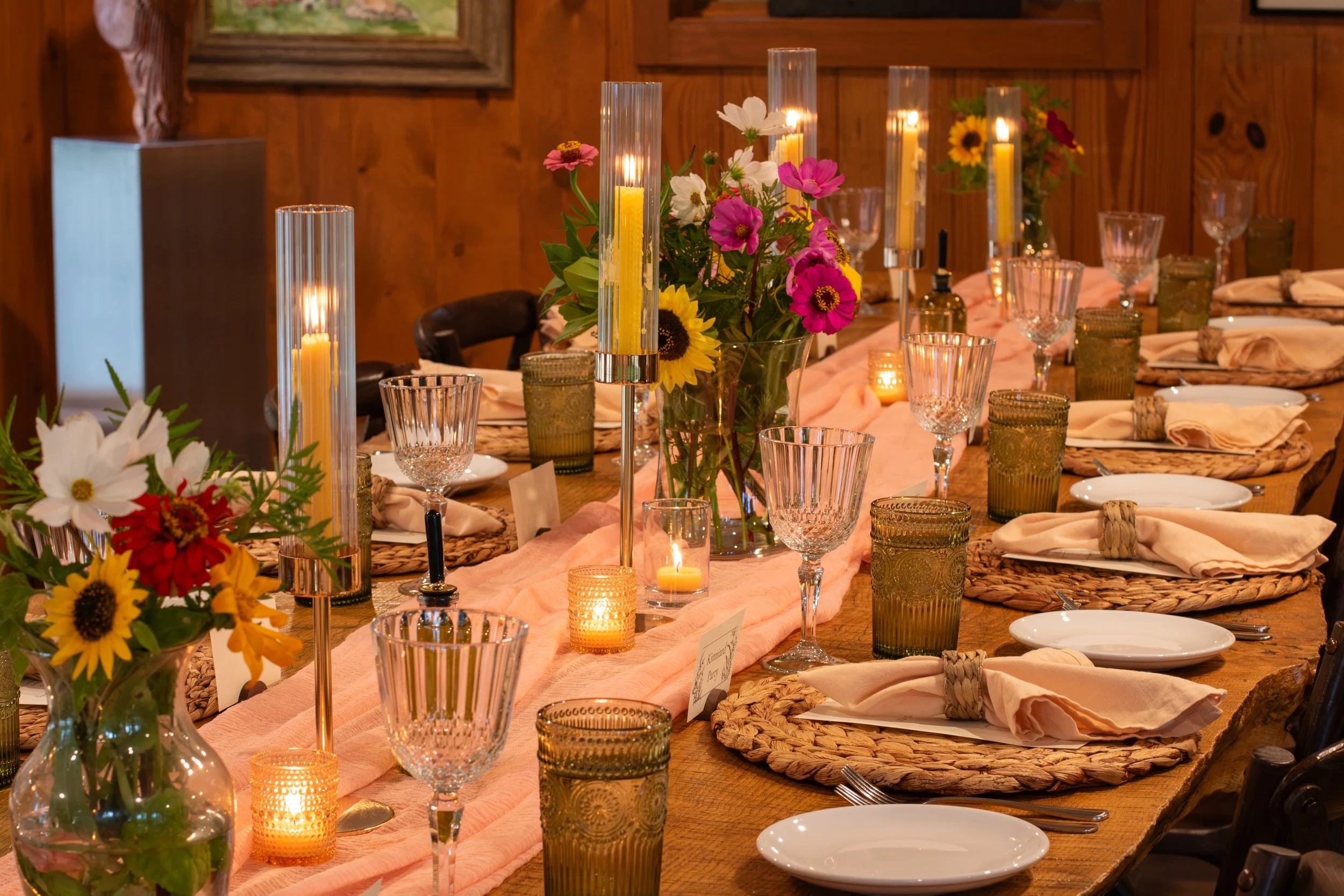 A beautifully decorated rustic dining table with a pink table runner, floral centerpieces featuring sunflowers, daisies, and pink flowers, surrounded by glassware, candles, and cloth napkins.