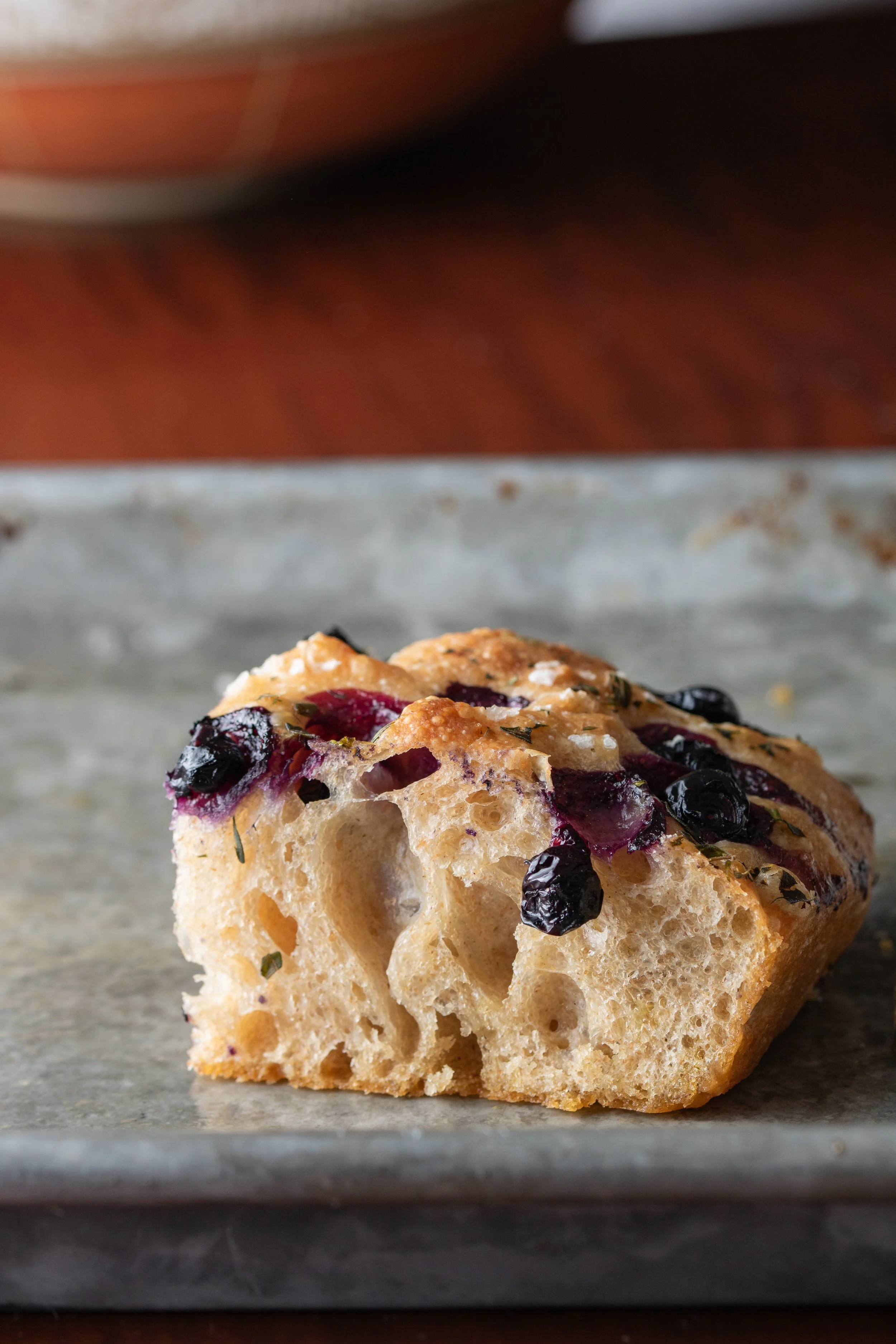Close-up of a piece of blueberry muffin with blueberries and herbs on top, on a gray surface, with a blurred background.