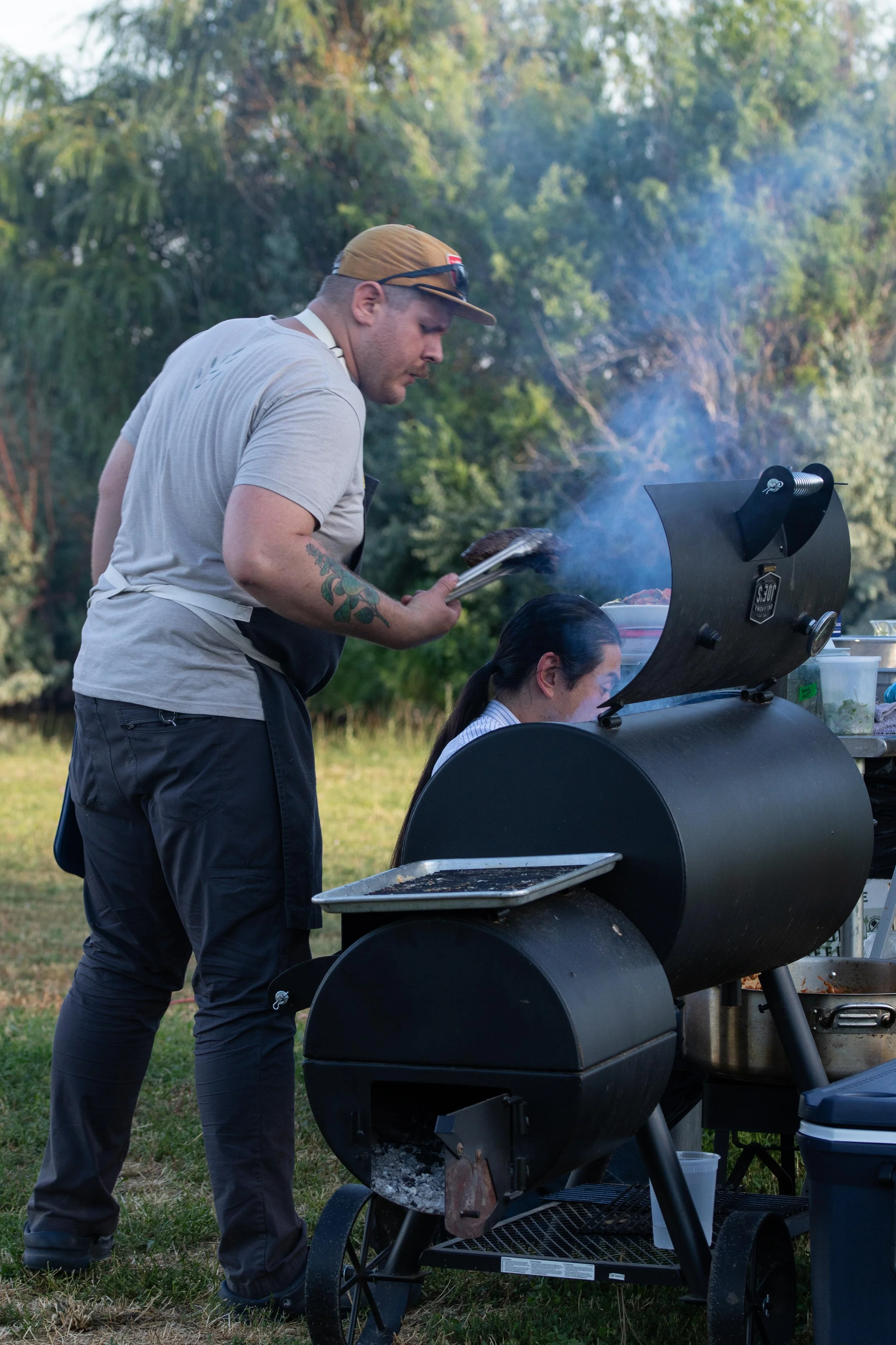 A man wearing a beige cap, gray t-shirt, and black apron is looking into a smoker grill while a woman with black hair tied back works nearby during an outdoor gathering in a grassy area surrounded by trees.