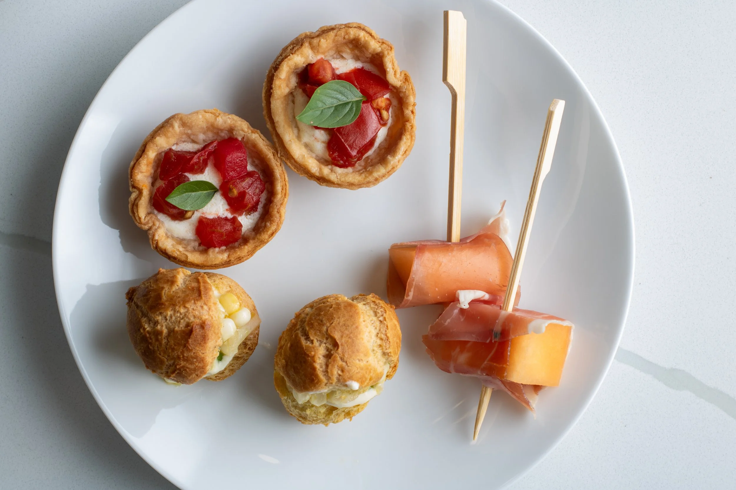 Plate with two fried cheese-filled appetizers topped with tomato and basil, a breaded and filled pastry, and slices of prosciutto-wrapped cheese on skewers.
