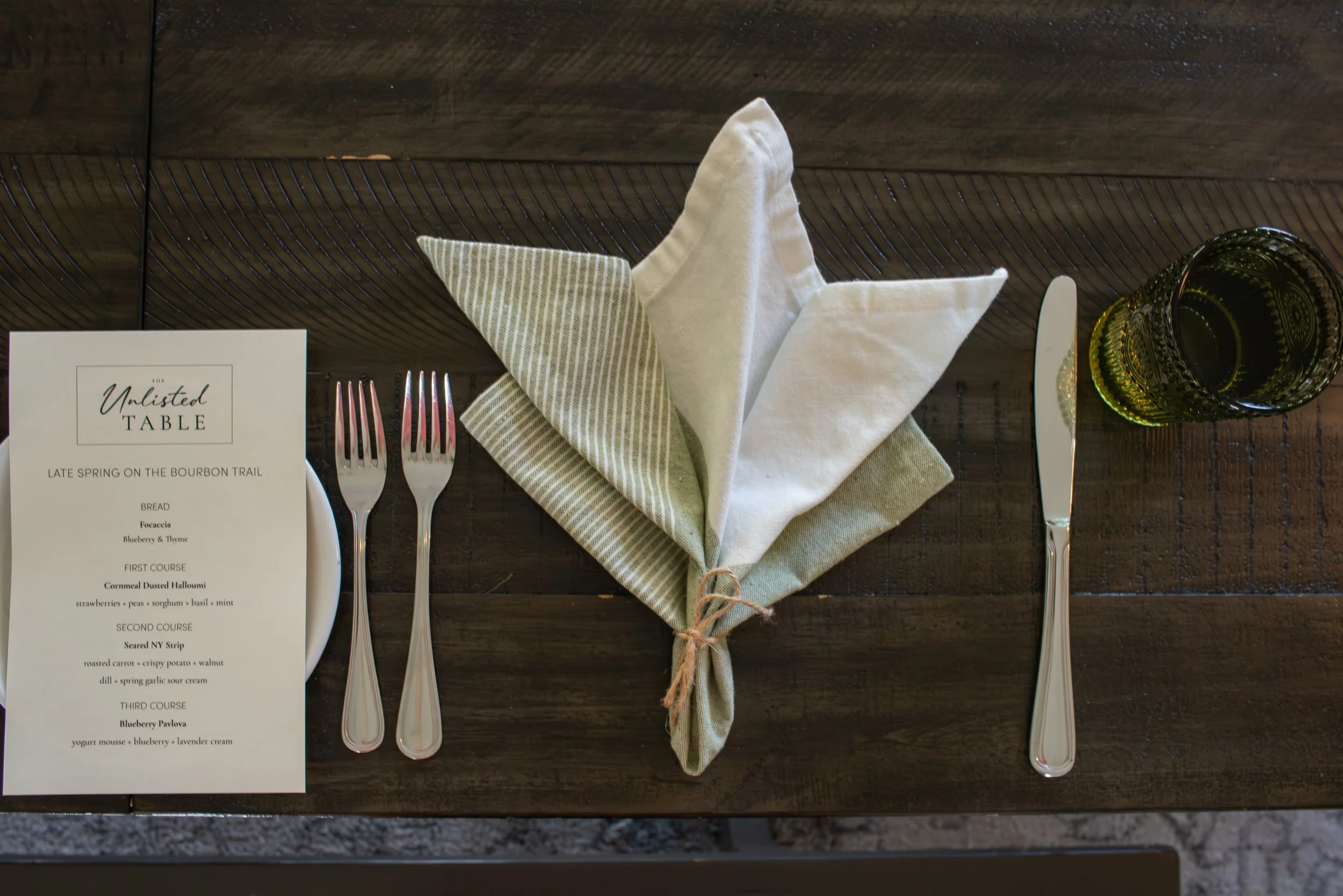 A place setting with a menu, a fork and knife, a cloth napkin folded into a fan shape, and a small glass bowl on a dark wooden table.