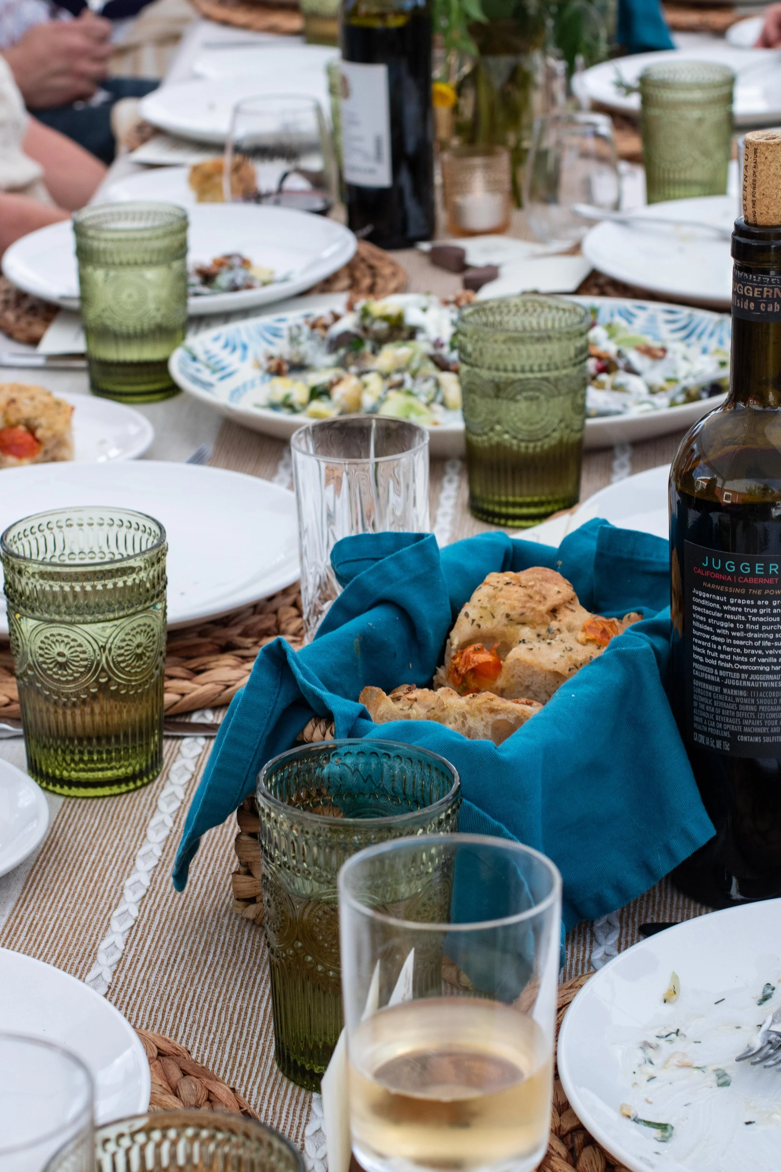 A dining table set for a meal with wine glasses, green textured drinking glasses, plates with food, a basket of bread, and a bottle of wine.