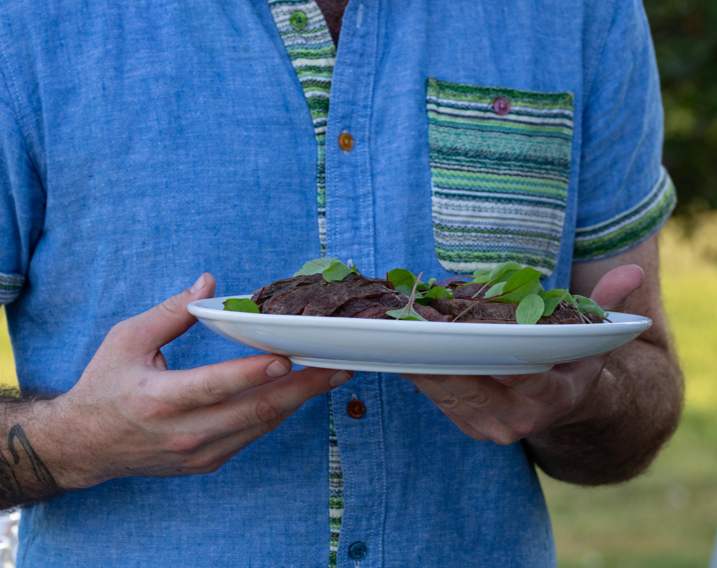 A person in a blue shirt holding a white plate with cooked meat and garnished with greens.