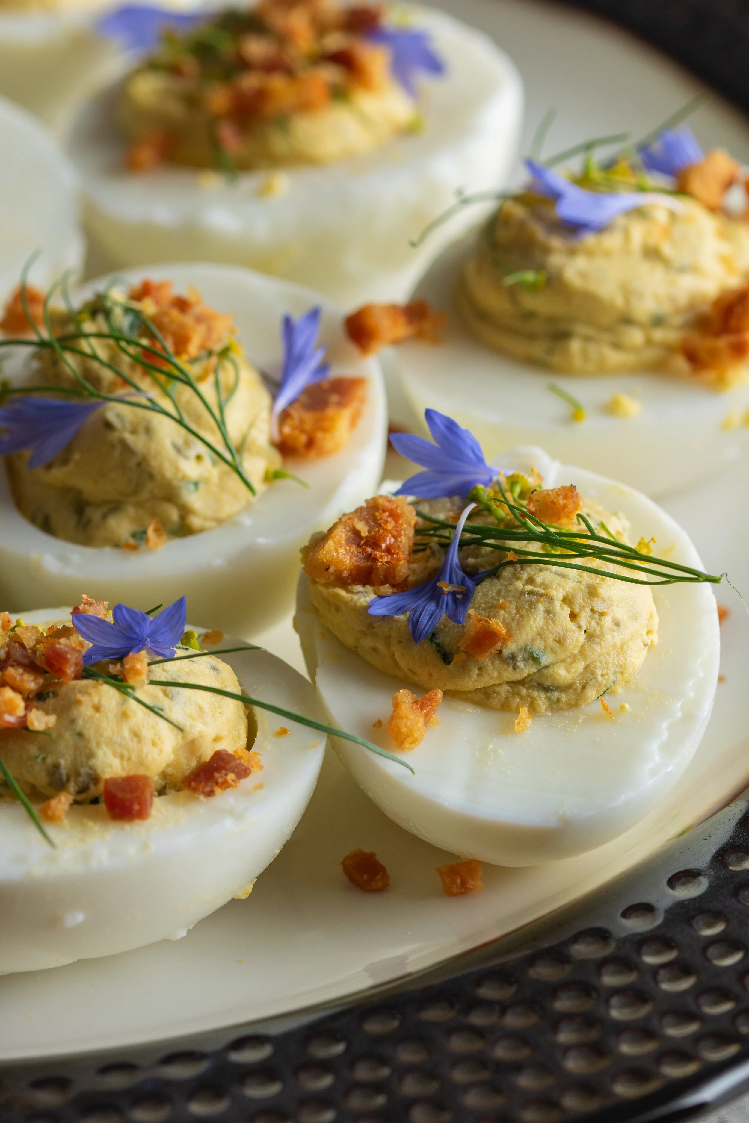 Close-up of halved hard-boiled eggs topped with egg salad, edible blue flowers, and crispy bacon bits on a white plate.