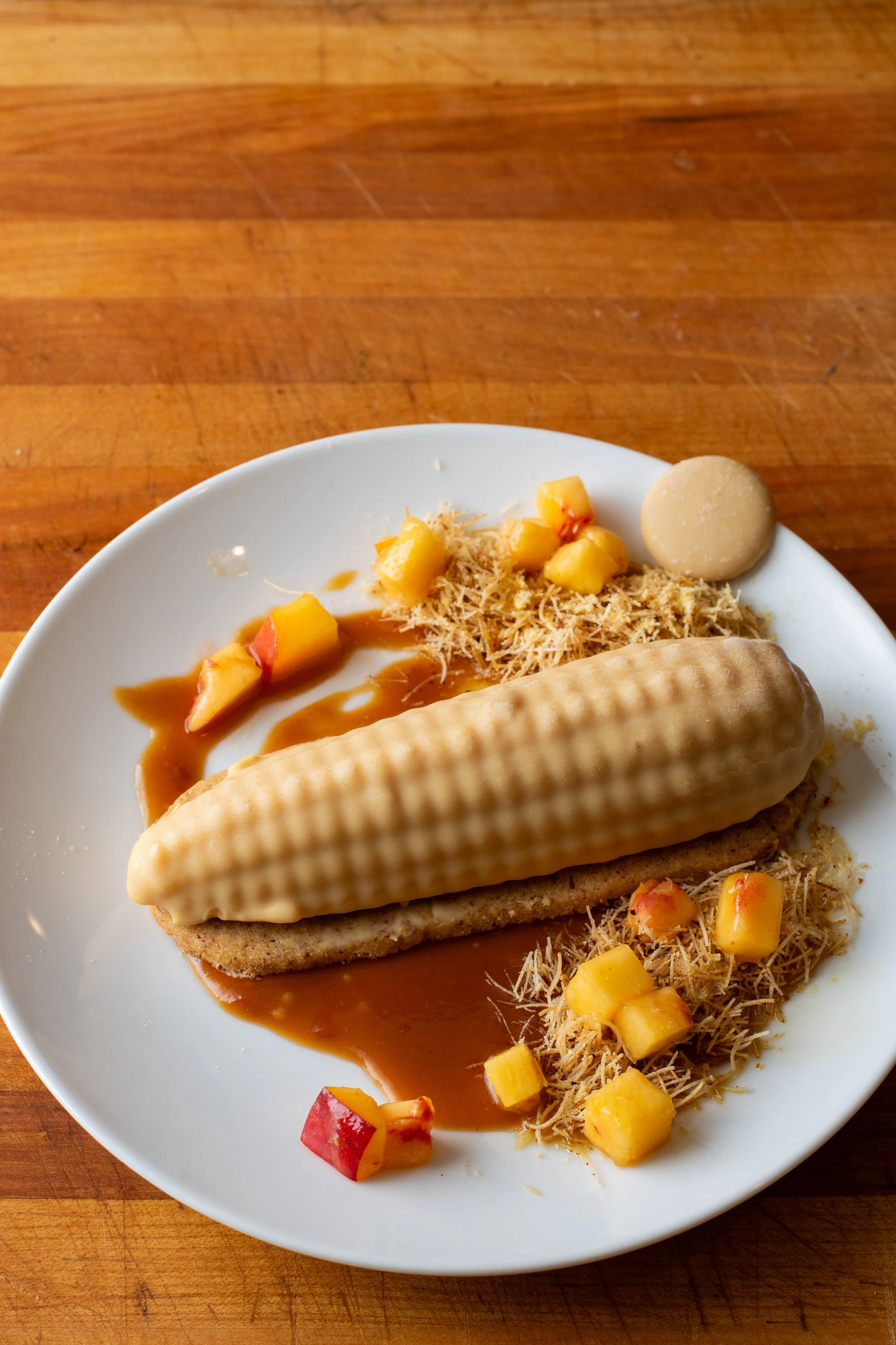A white plate with a dessert topped with a wafer, surrounded by diced peach and apple pieces, with crushed graham crackers and caramel sauce, set on a wooden surface.