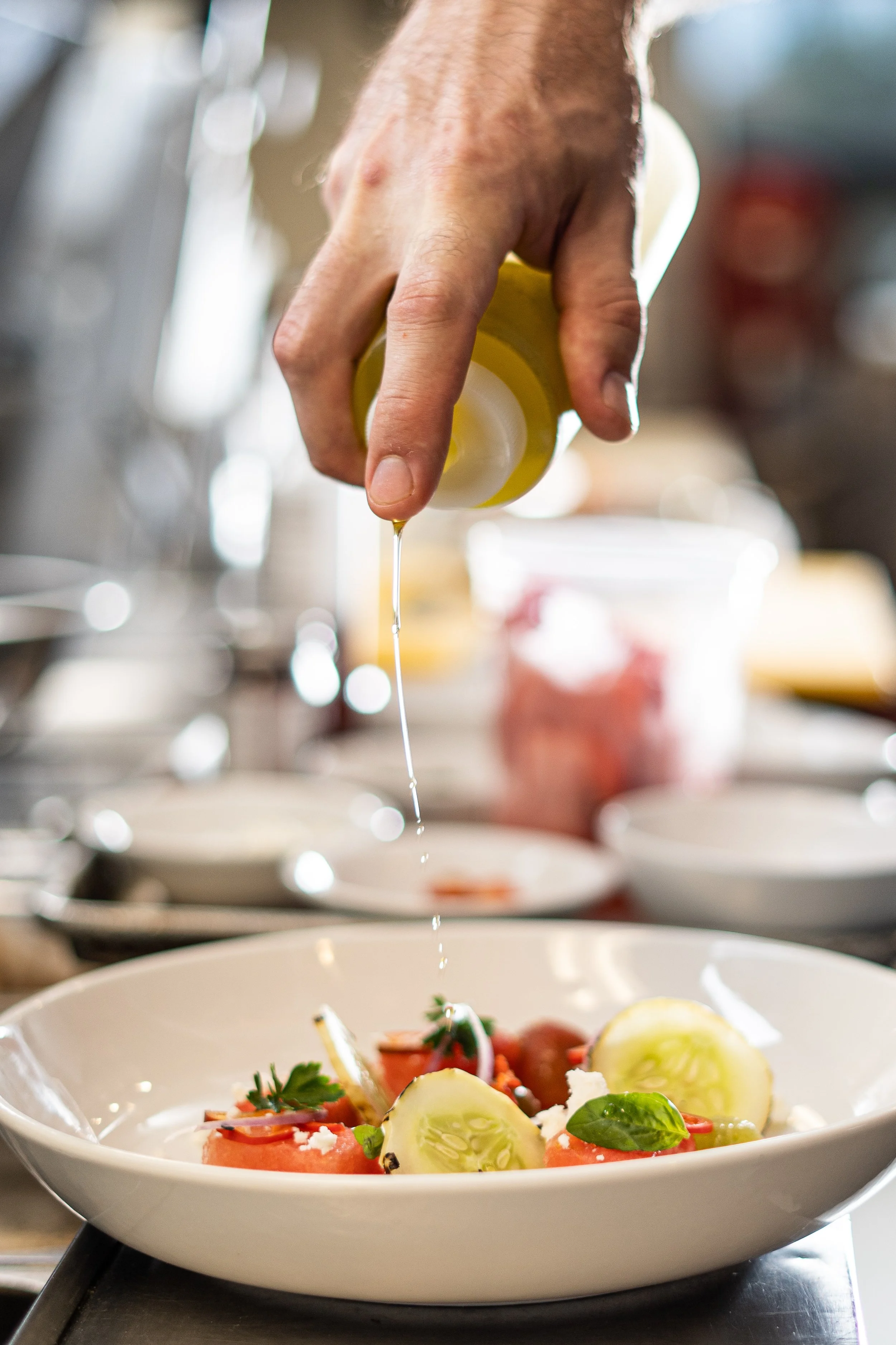 A chef drizzling dressing over a plate of fresh salad with tomatoes, cucumbers, and herbs.