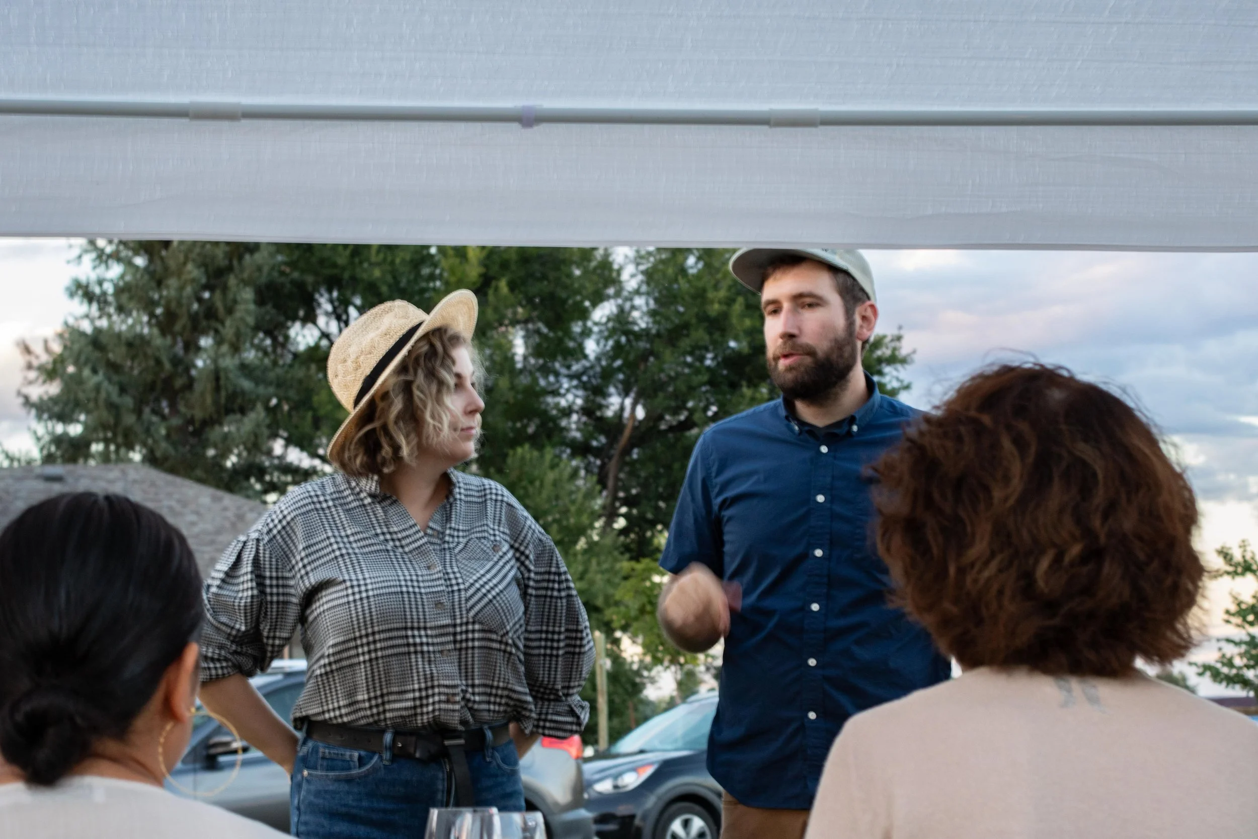 A group of people gathered outdoors, with two individuals standing and speaking to three seated women, during what appears to be an outdoor event or meeting in the evening.