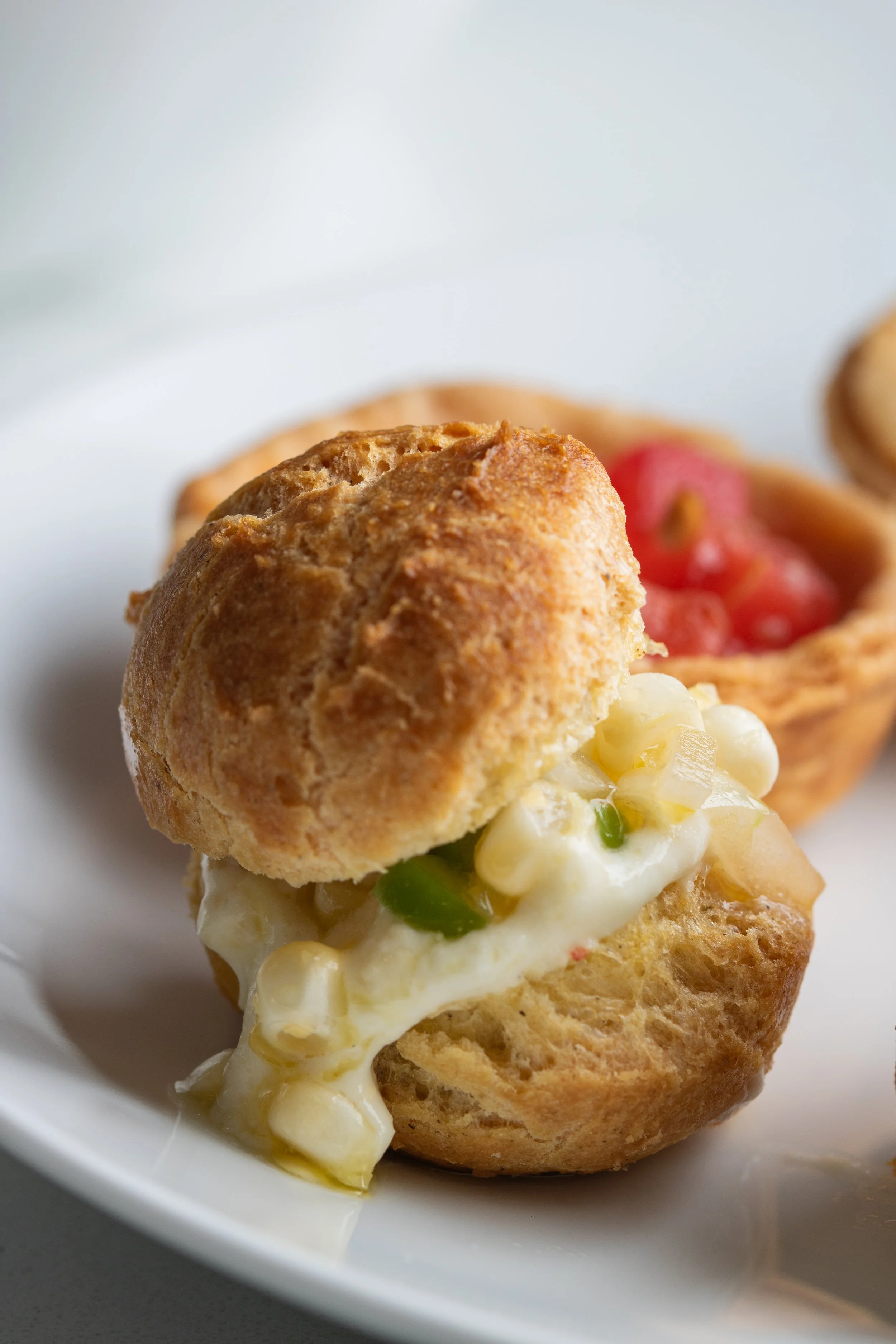 Close-up of a savory pastry with a golden-brown exterior, filled with creamy cheese, corn, and green herbs, on a white dish.