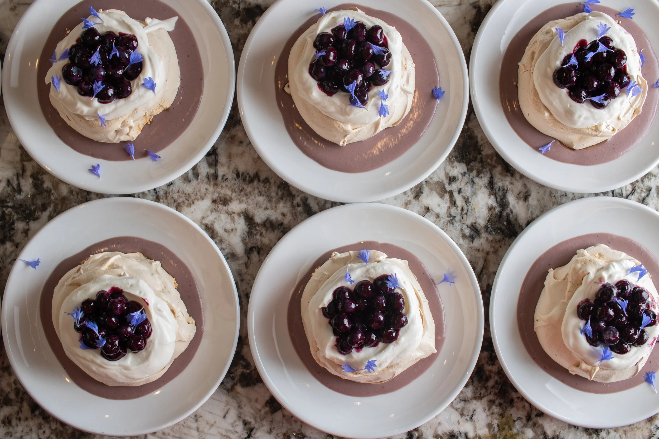 A row of individual pavlova desserts topped with blueberries and edible blue flowers on white plates, displayed on a granite countertop.