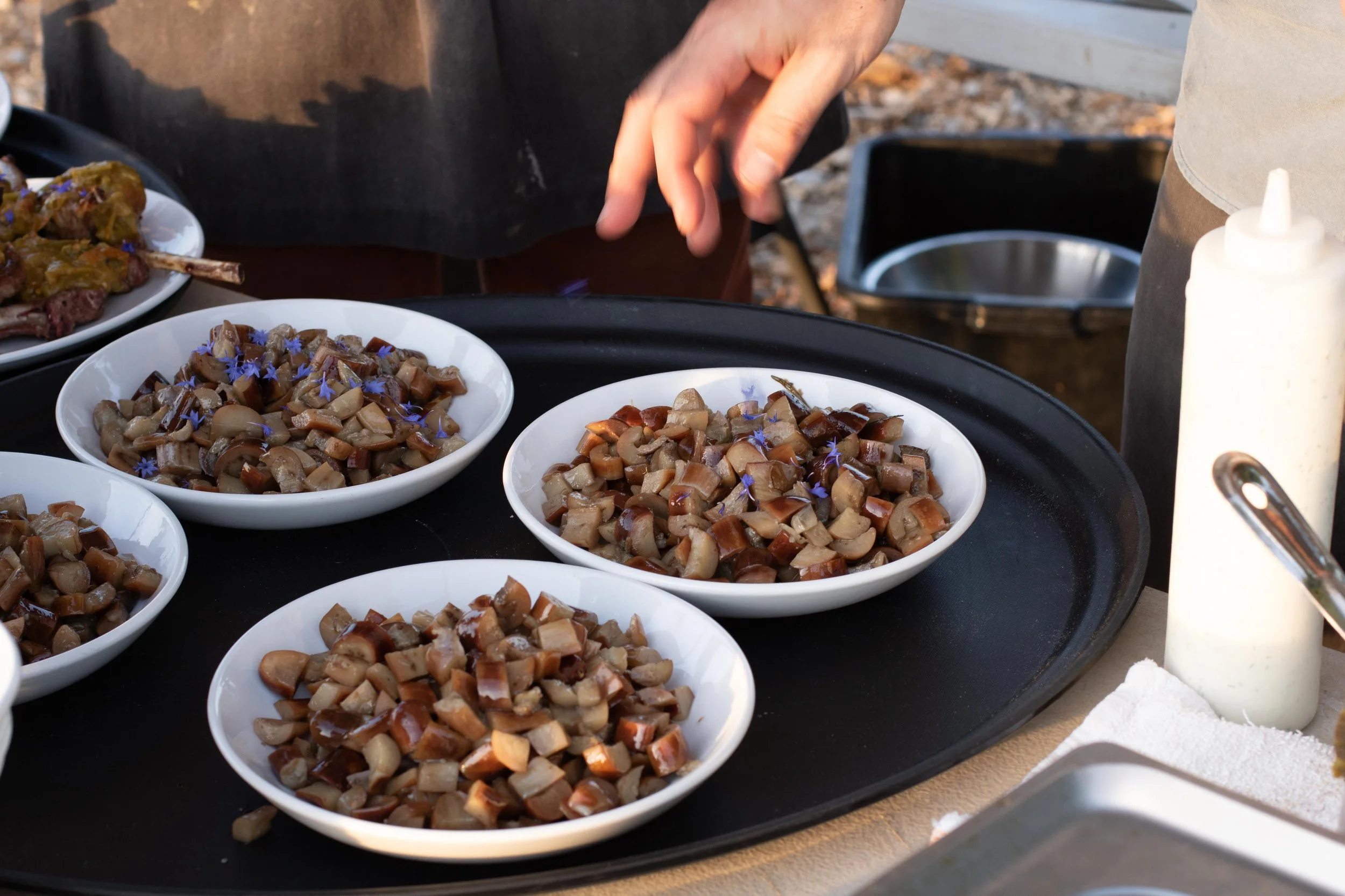 Bowls of chopped cooked mushrooms garnished with edible flowers on a black tray at a food stand.