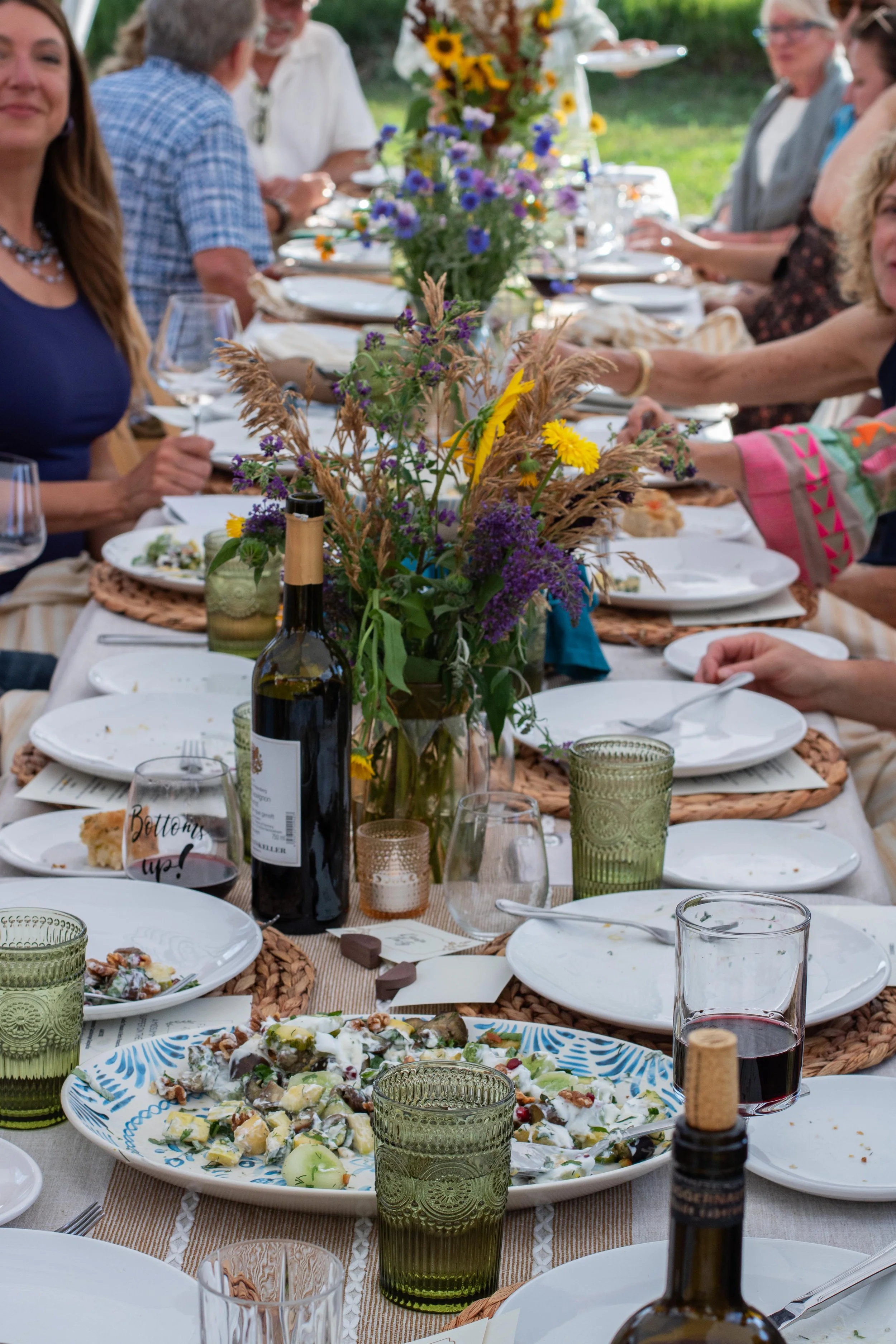 A large outdoor dinner table set with plates, glasses, and a centerpiece of wildflowers. The table has some remaining food, a bottle of wine, and decorative items, with people sitting around it in a garden setting.