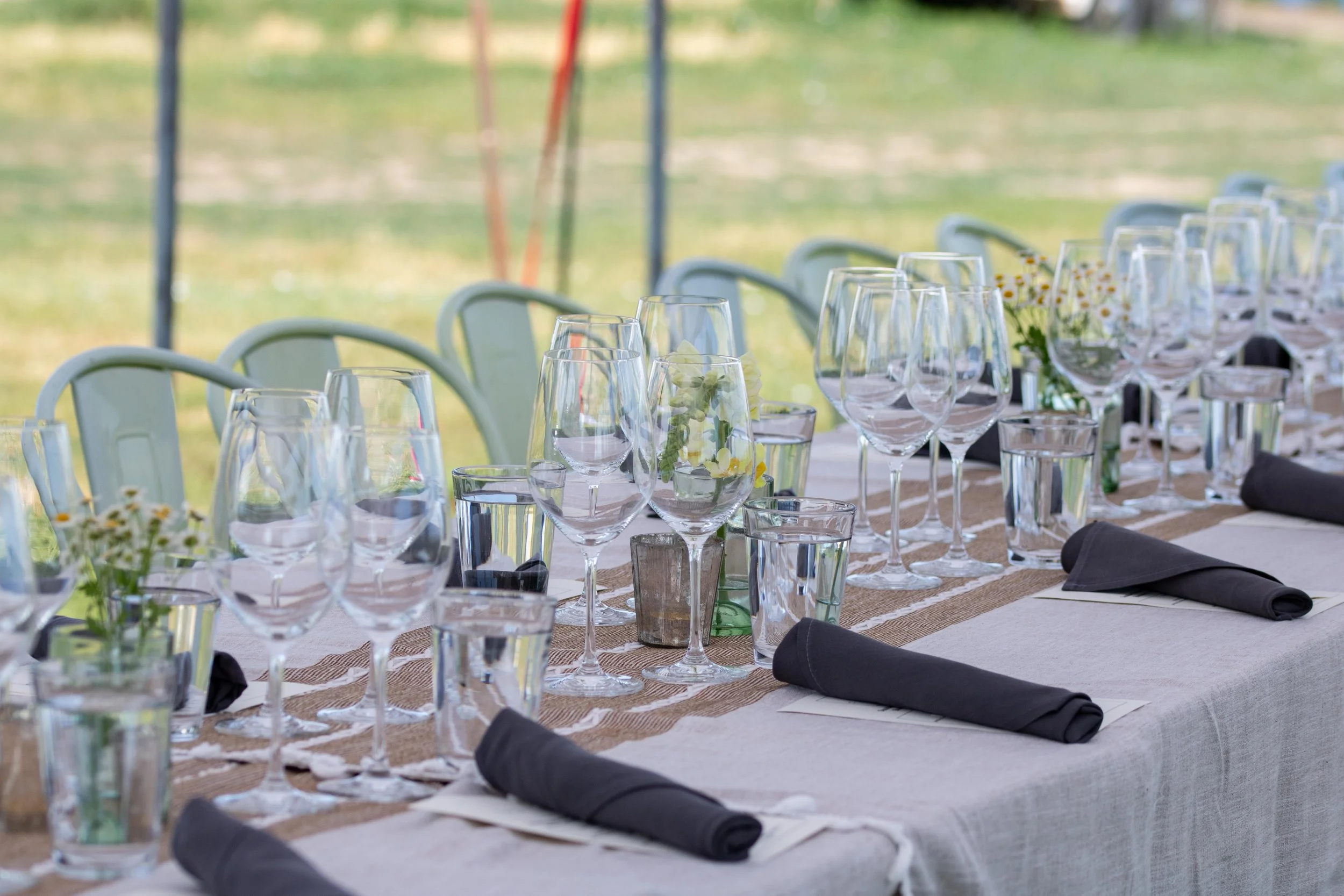 A long outdoor table set for a formal event with wine glasses, water glasses, black napkins, and small floral centerpieces, with green chairs in the background.