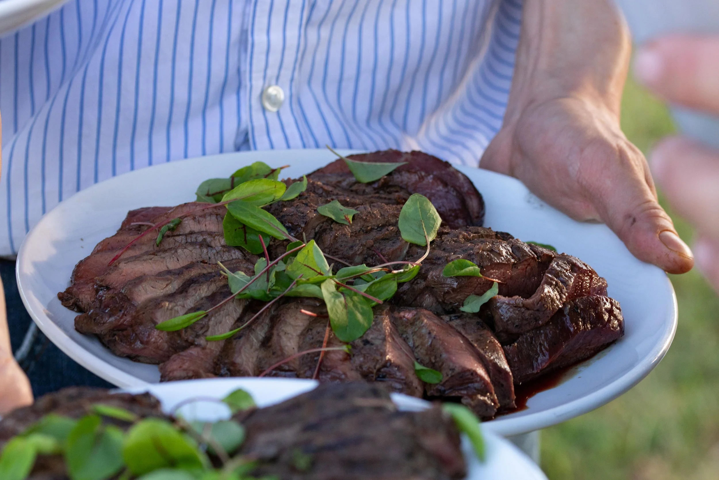 Person holding a white plate with cooked steaks garnished with green herbs.