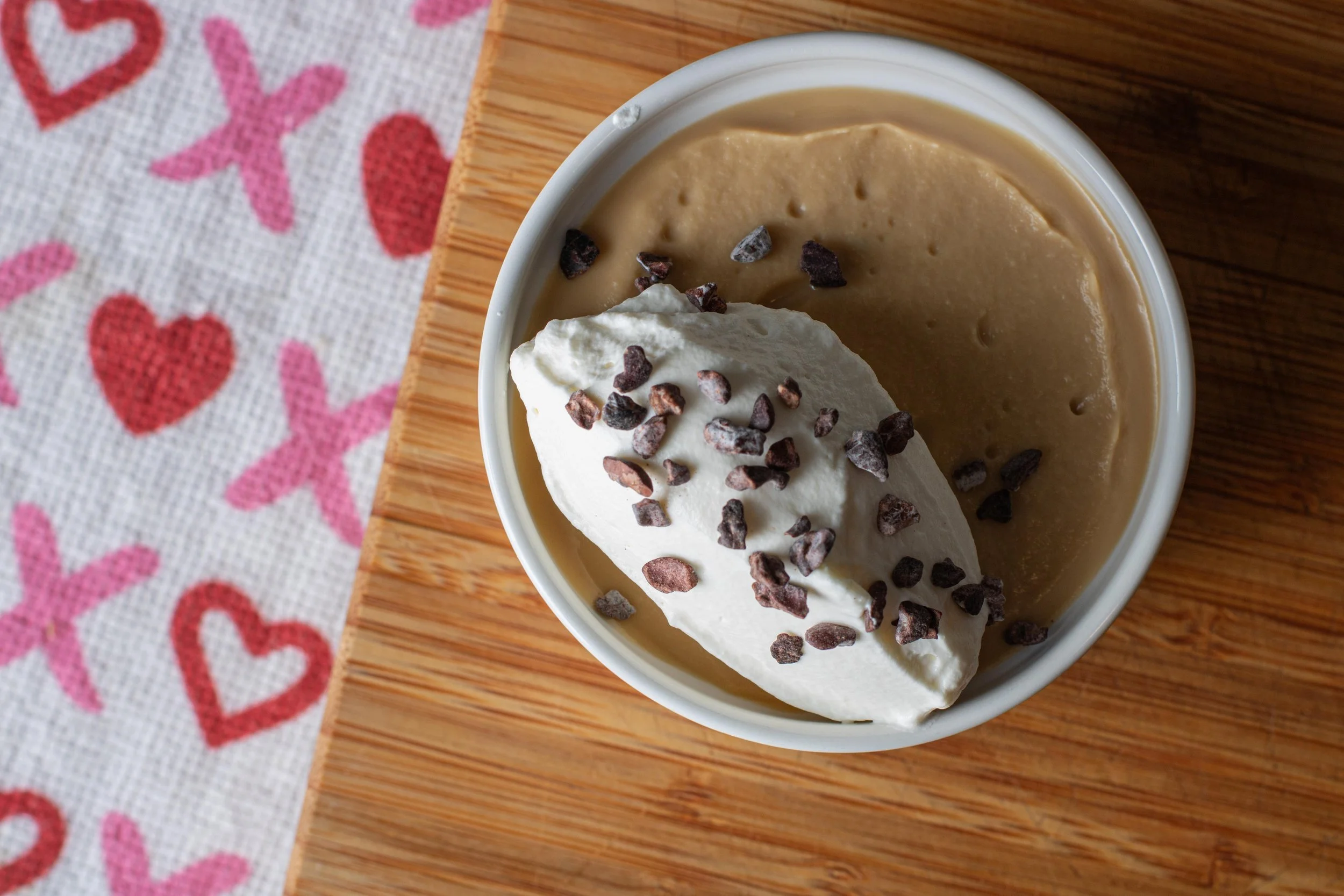 Overhead shot of a white ramekin filled with pudding and topped with a whipped cream quenelle and cocoa nibs