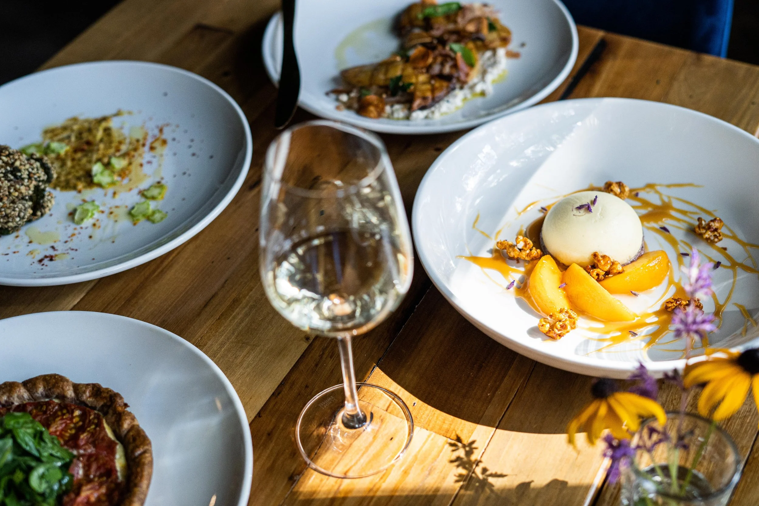 A dining table with various dishes including a white plate with a dessert garnished with peaches, caramel sauce, walnuts, and edible flowers, a glass of white wine, and other plates with assorted foods in the background.