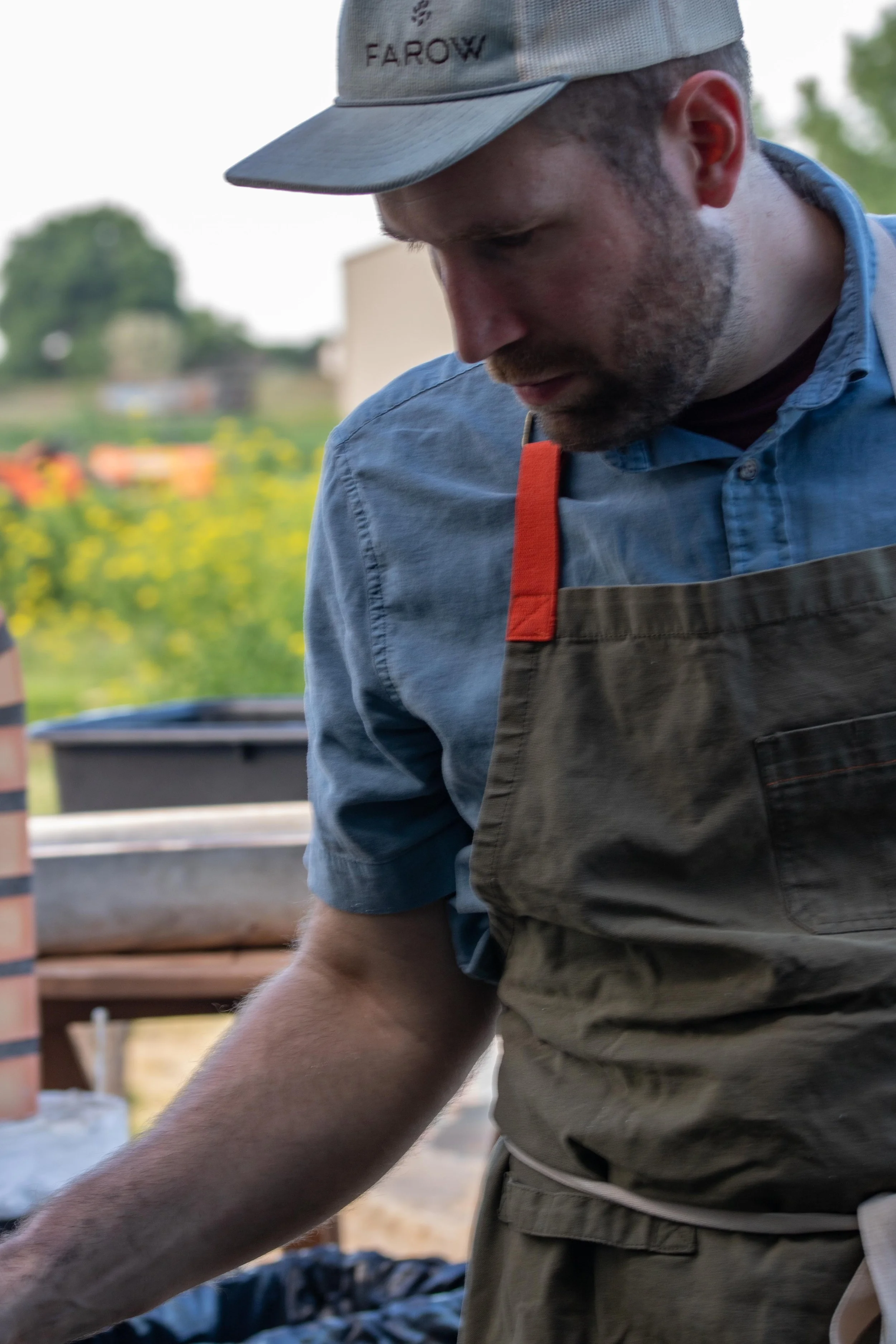 A man wearing a blue denim shirt, an apron, and a gray cap, looking down while cooking outdoors at a grill with a blurred background of yellow flowers and greenery.