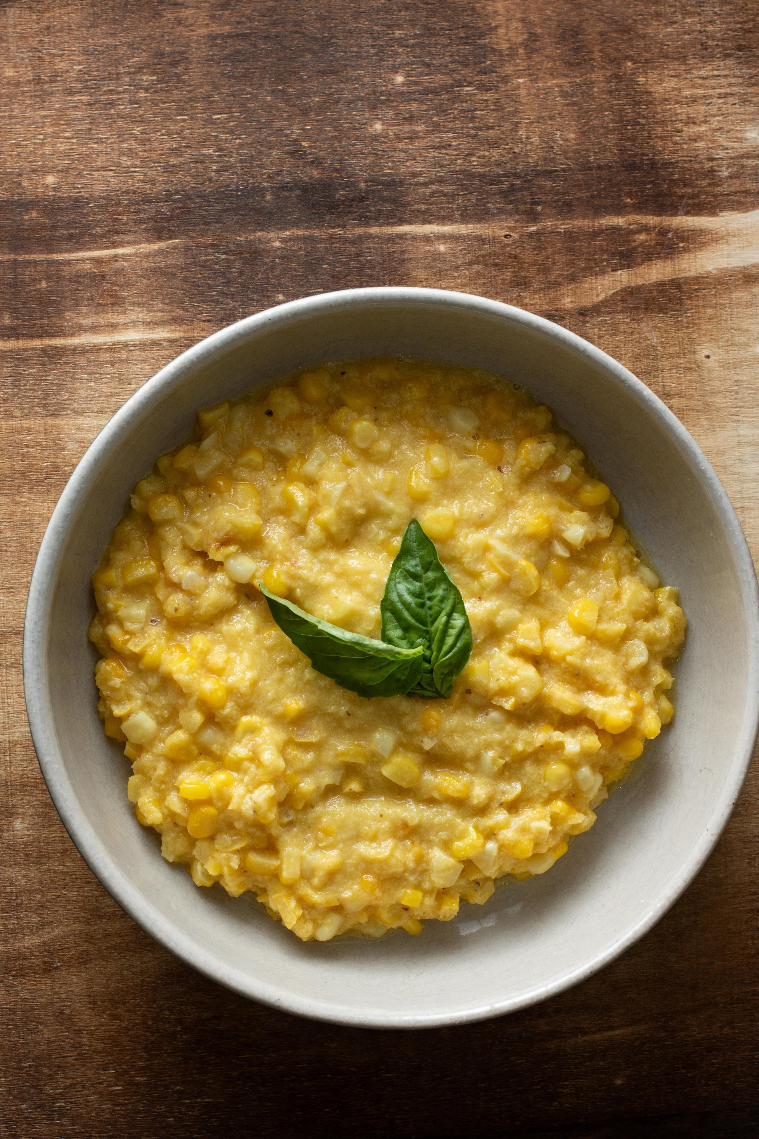A bowl of creamed corn garnished with fresh basil leaves on a wooden surface.