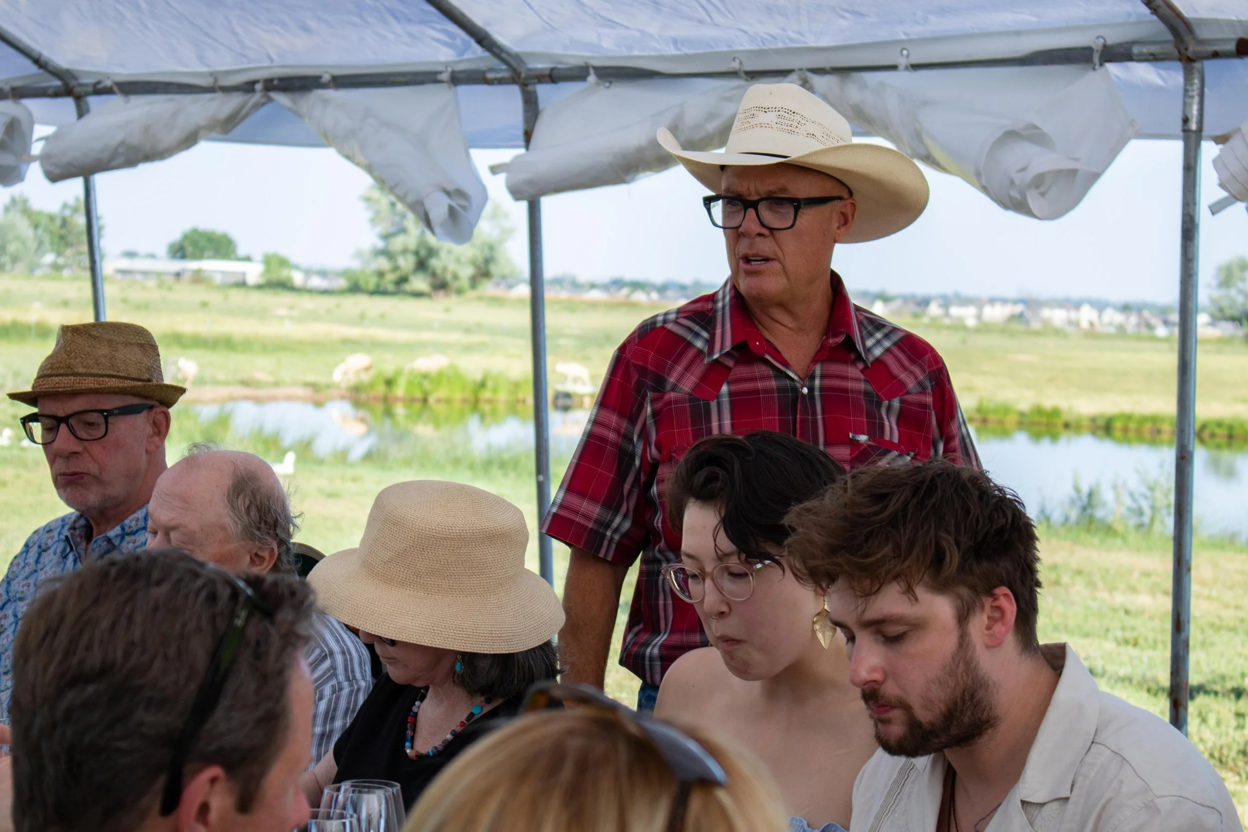 A group of people sitting outdoors at a table, with a man in a red plaid shirt and white cowboy hat standing and looking towards the side, surrounded by others with hats and glasses, with a scenic landscape of water and fields in the background.