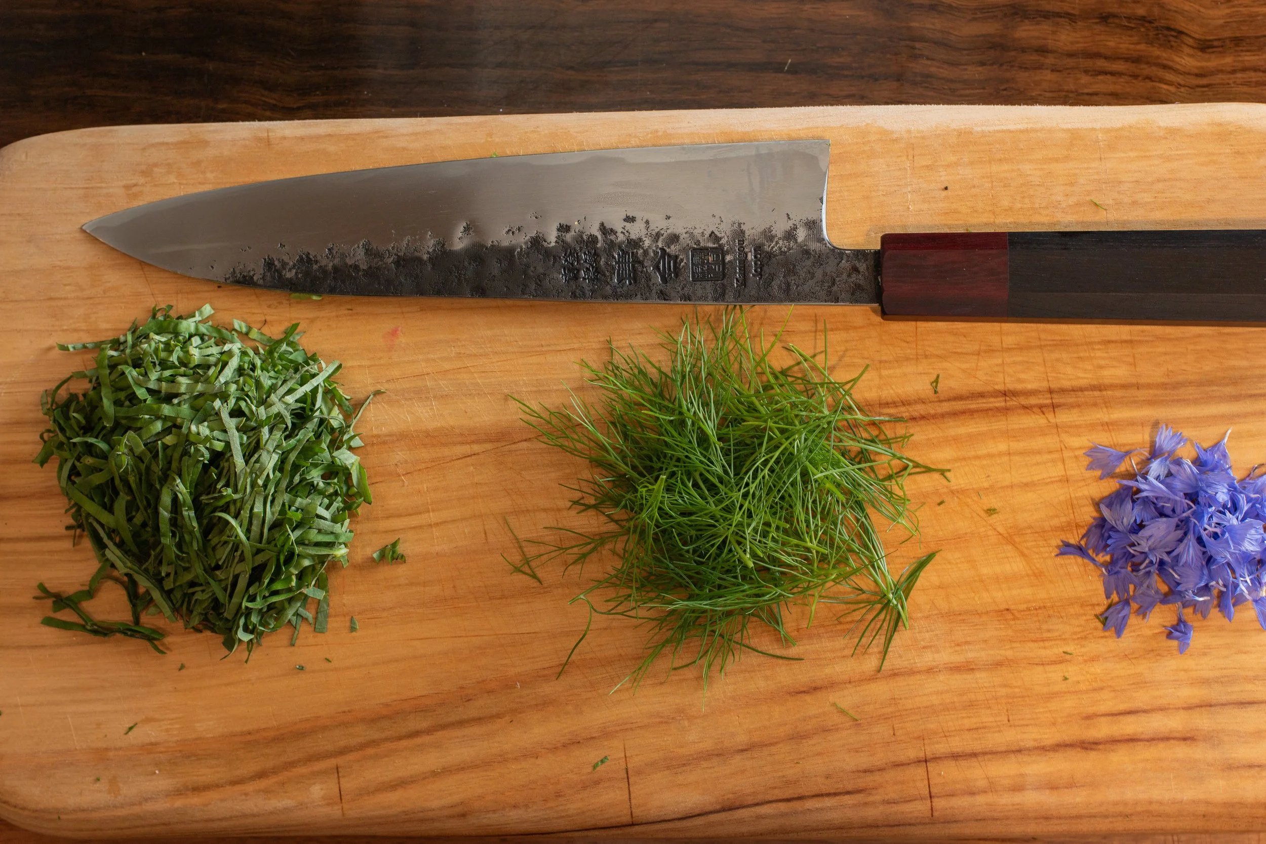 A kitchen knife resting on a wooden cutting board with chopped green herbs and purple flowers nearby.