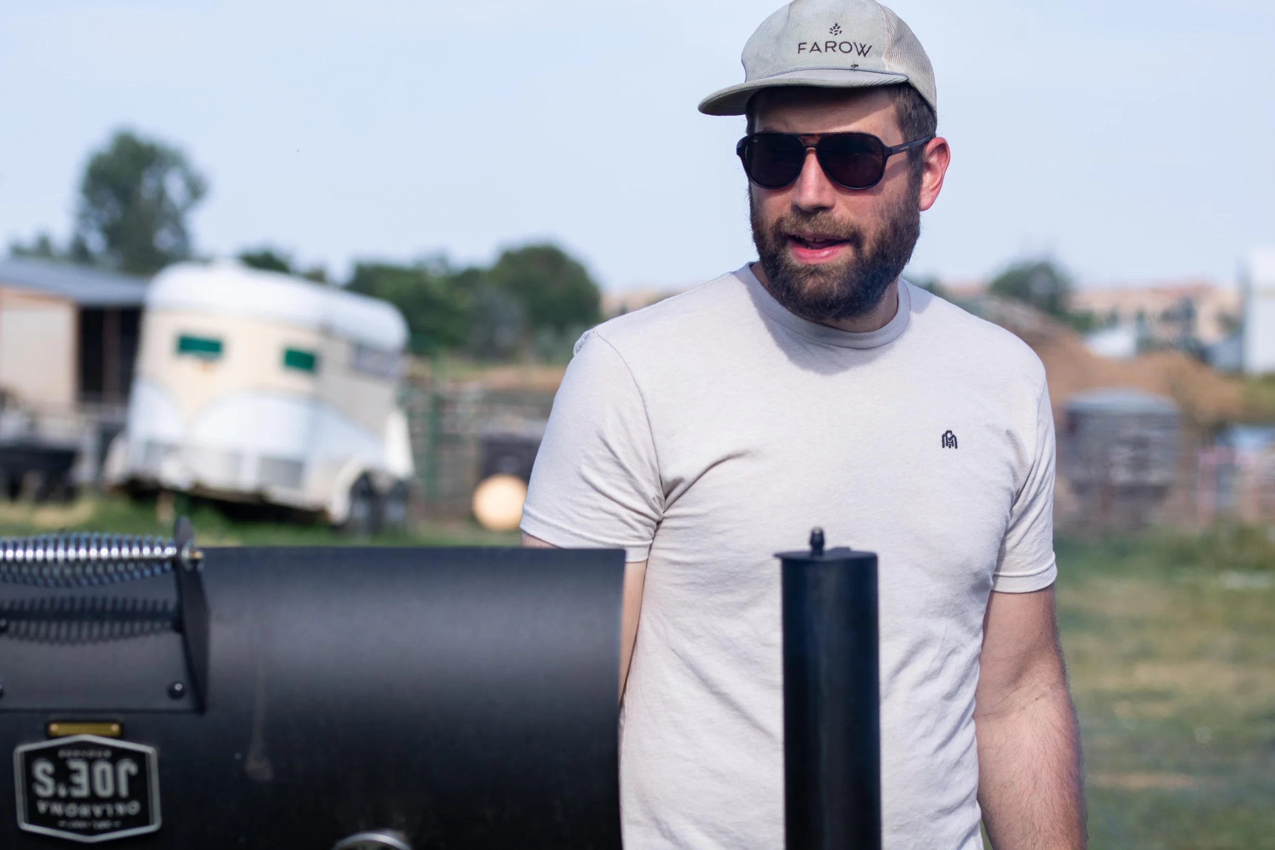 A man with a beard wearing sunglasses, a light gray cap, and a gray t-shirt standing outdoors with a barbecue grill in front of him, with a trailer and trees in the background.