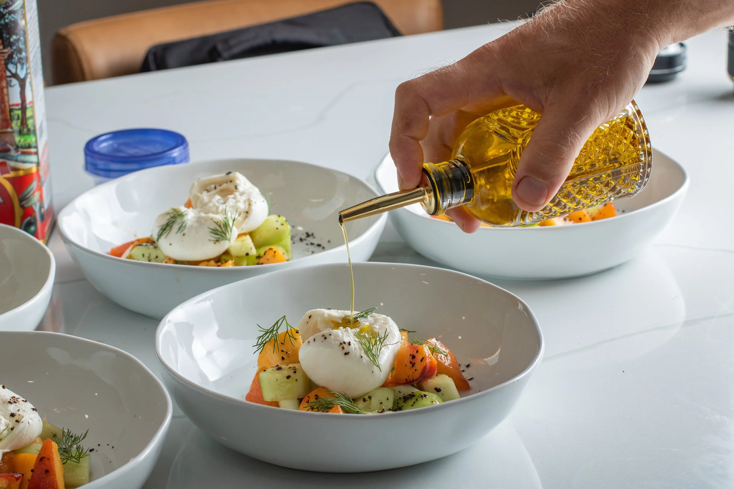 Person pouring olive oil over a bowl of diced melon and mozzarella garnished with herbs. Additional bowls of similar salads are visible on a white table.