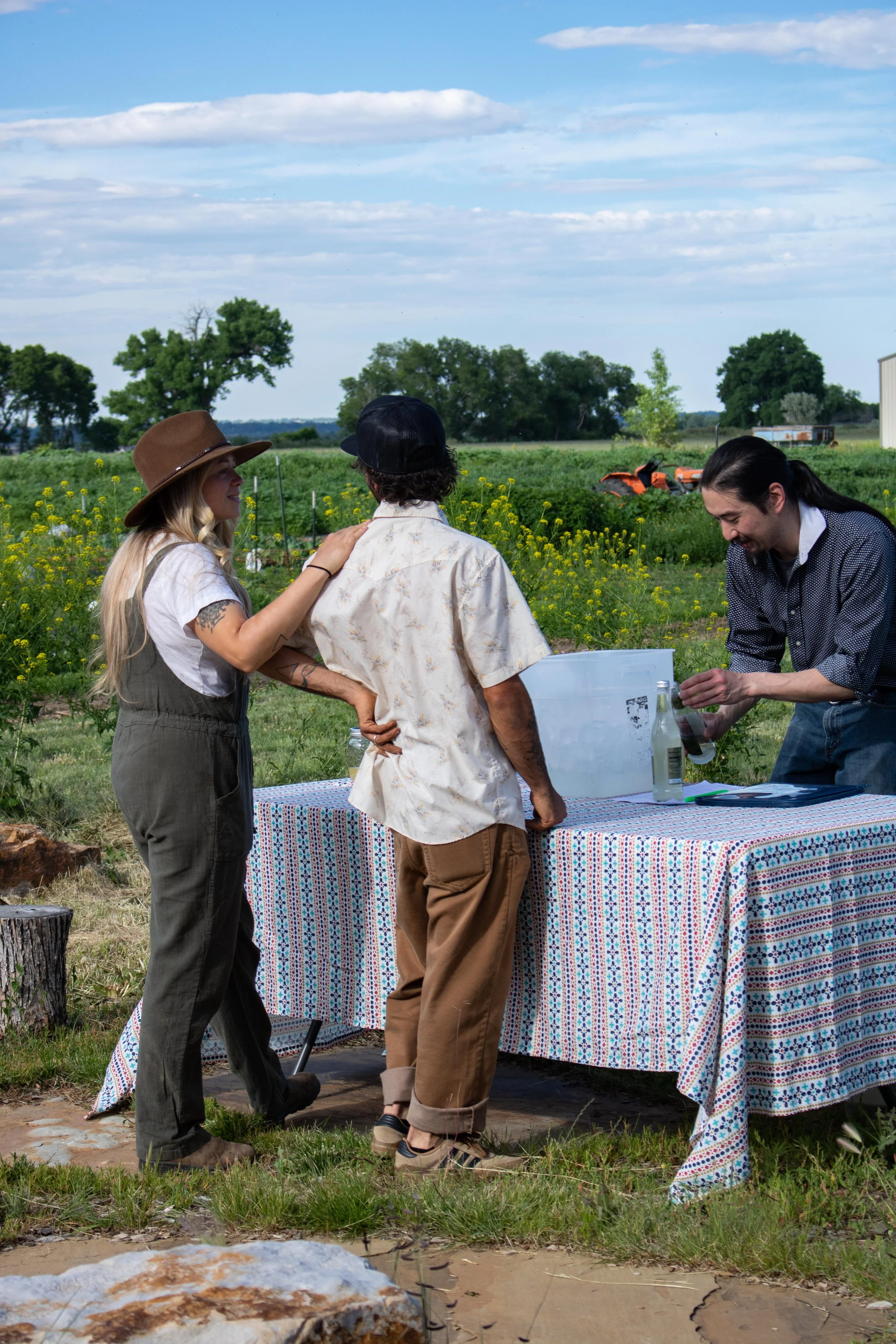 Two women and one man standing outdoors near a table with a colorful cloth, with farm fields and trees in the background, engaging in conversation and laughing.