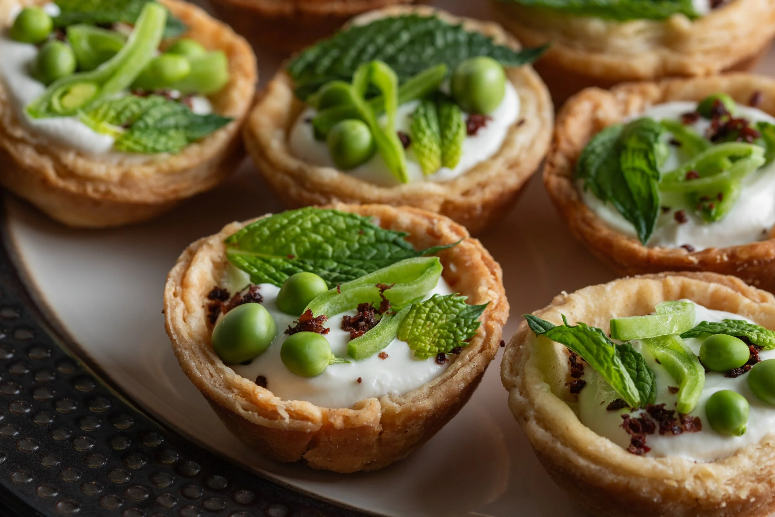 Close-up of savory tartlets topped with fresh mint, green peas, and creamy filling on a black textured tray.