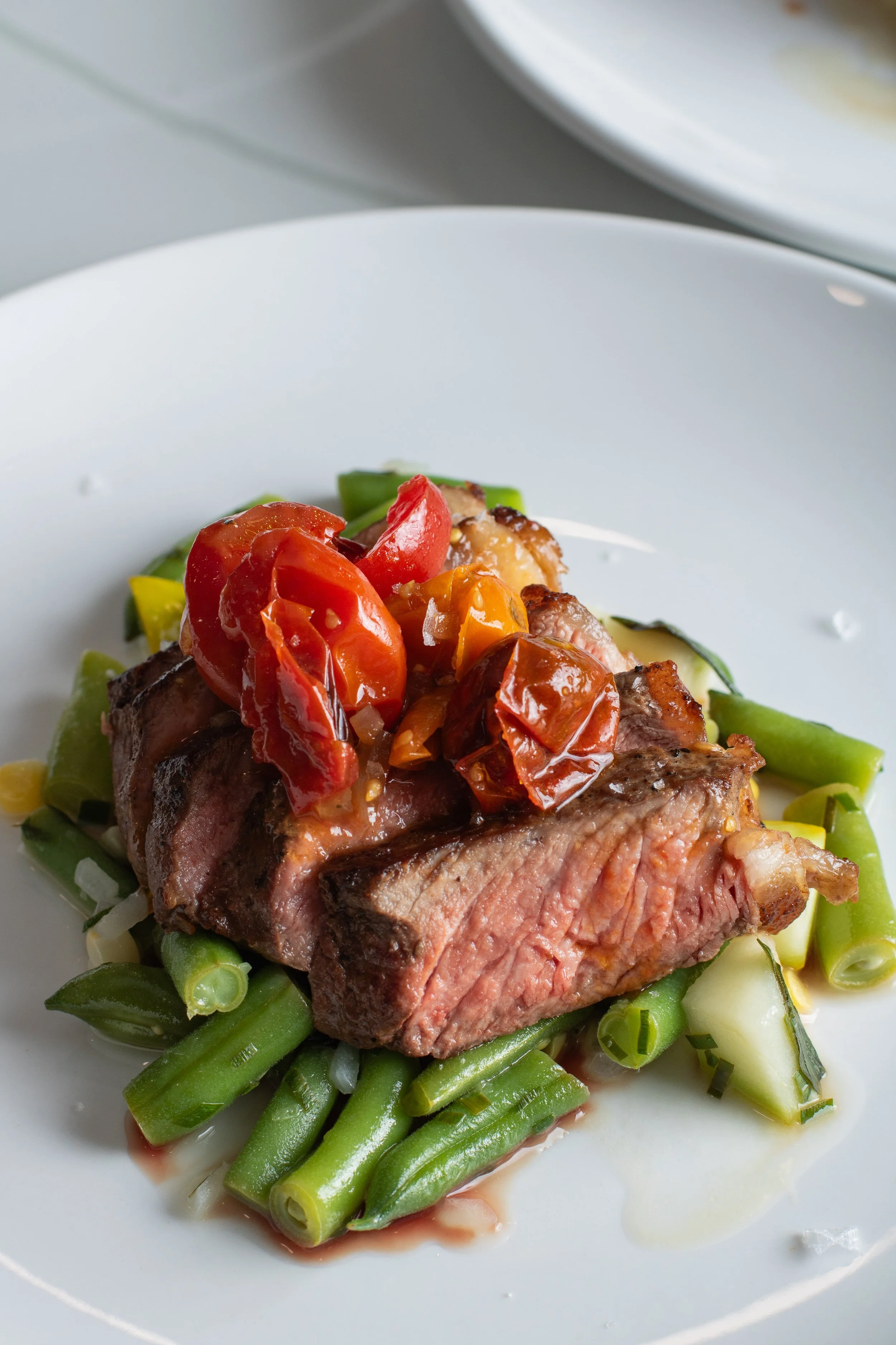 A close-up view of a cooked steak topped with tomatoes and onions, placed on a bed of green beans on a white plate.