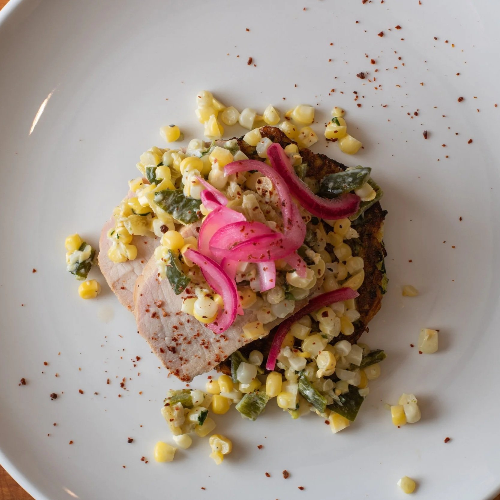 A plate with a piece of cooked meat topped with corn salad with pickled onions and chopped green herbs, and scattered corn and spices around on the plate.