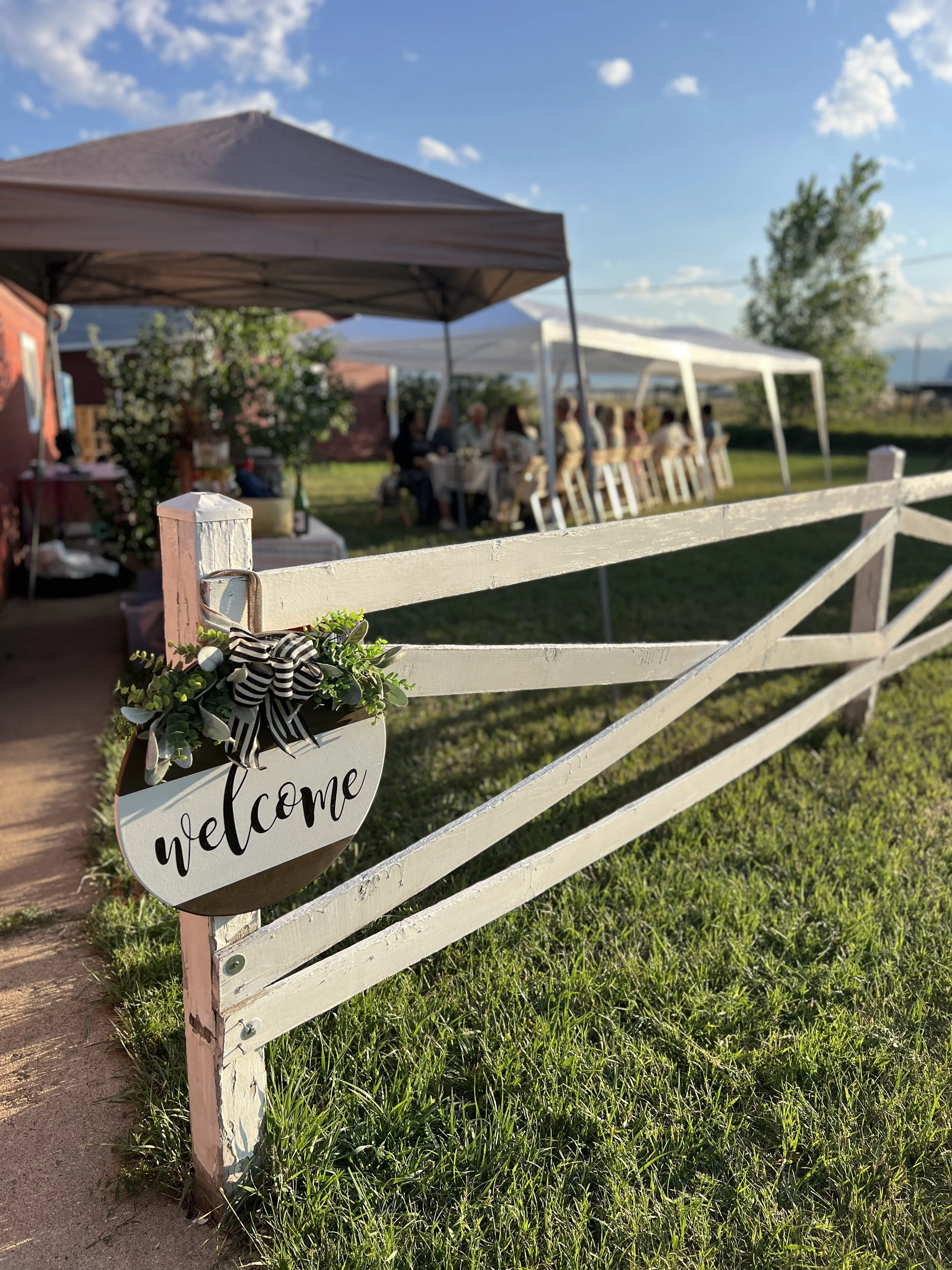 White wooden fence with a decorative 'welcome' sign and a black and white striped bow, leading to outdoor gathering under tents with people sitting at a long table, in a grassy area with a barn and trees, during a sunny day.