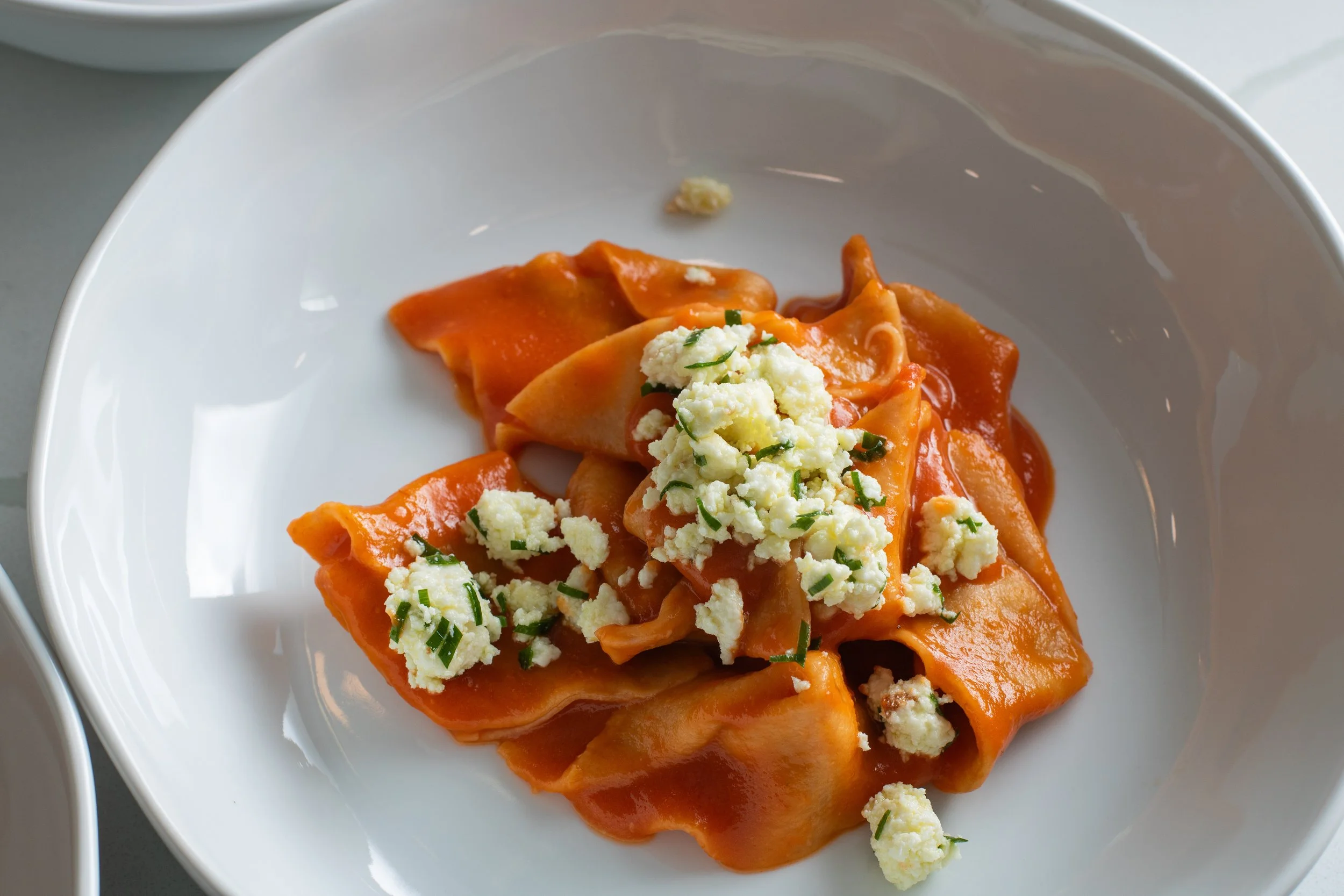 Close-up of cooked ravioli pasta with tomato sauce, topped with crumbled cheese and chopped herbs, on a white plate.