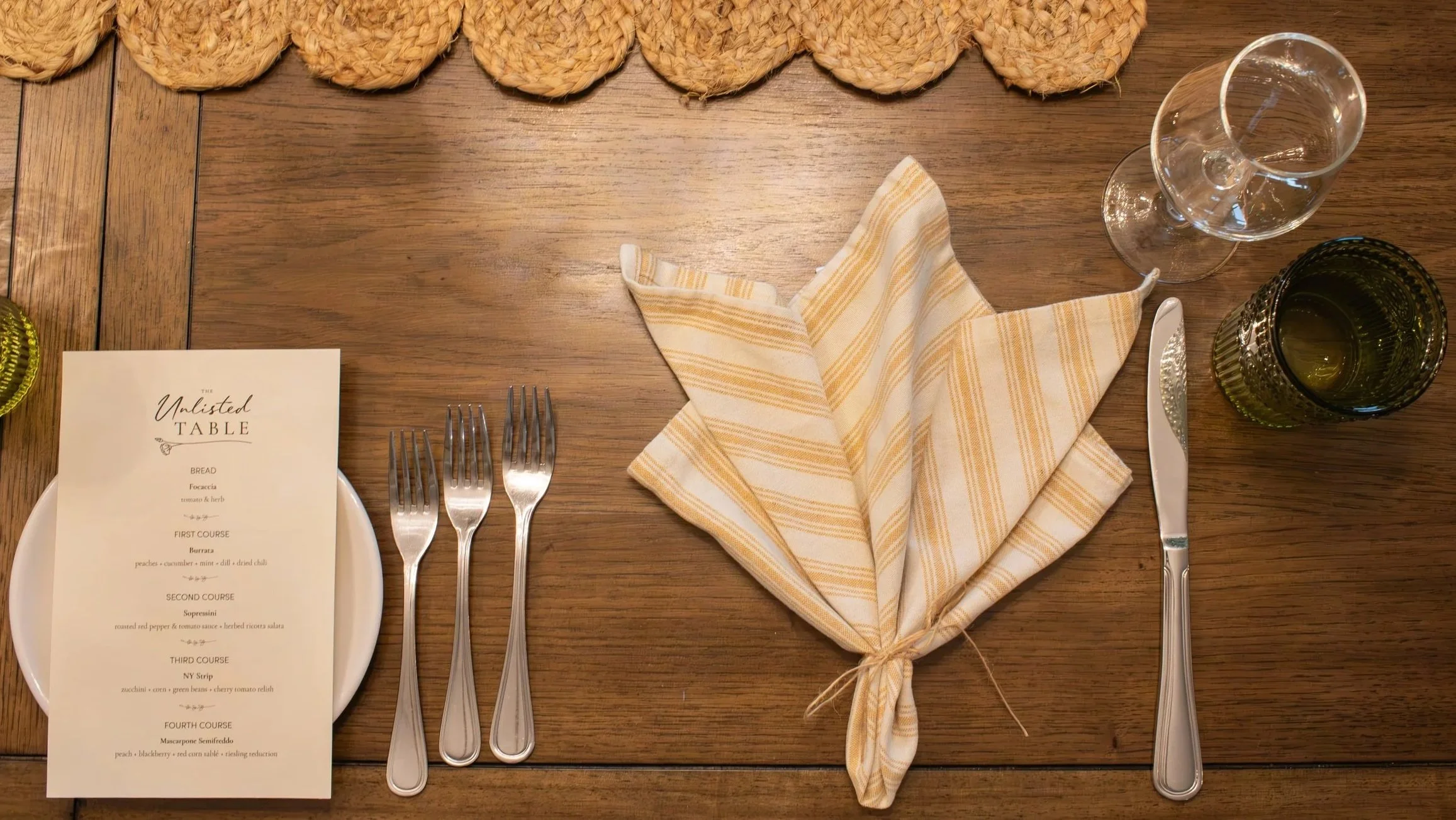 A dining table set with cutlery, a folded cloth napkin, glassware, and a printed menu, with woven placemats along the top edge.