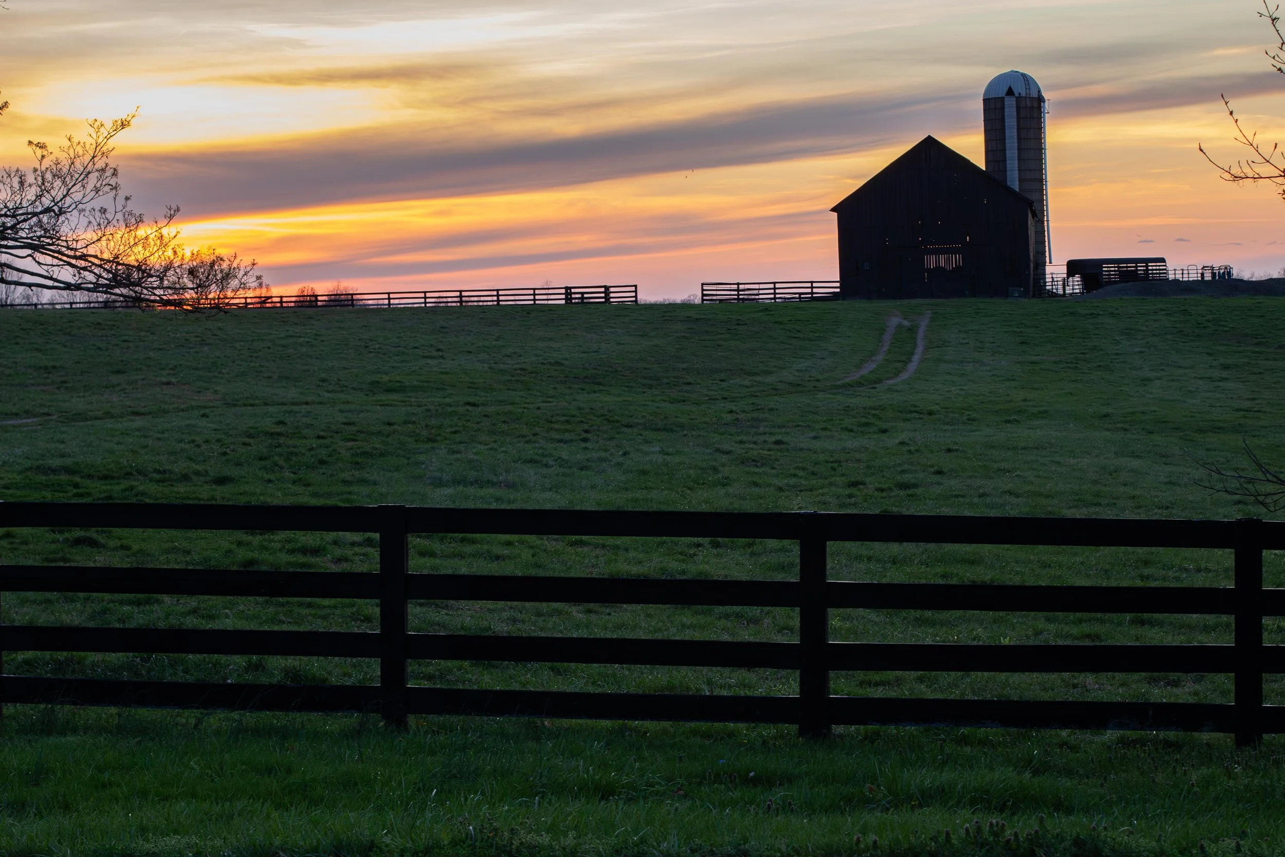 Sunset over a farm with a dark barn and silo, green grassy field, and a wooden fence in the foreground.