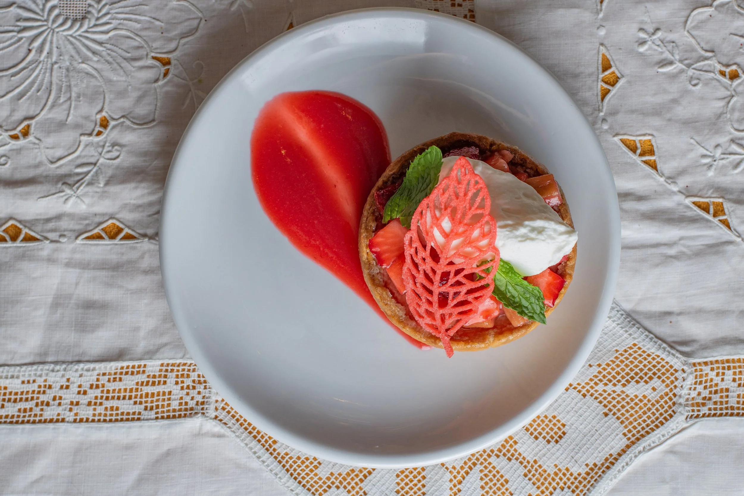 A dessert tart with strawberries, whipped cream, mint leaves, and a pink decorative pattern, served with a sliced fresh tomato on a white plate on a table with a embroidered tablecloth.