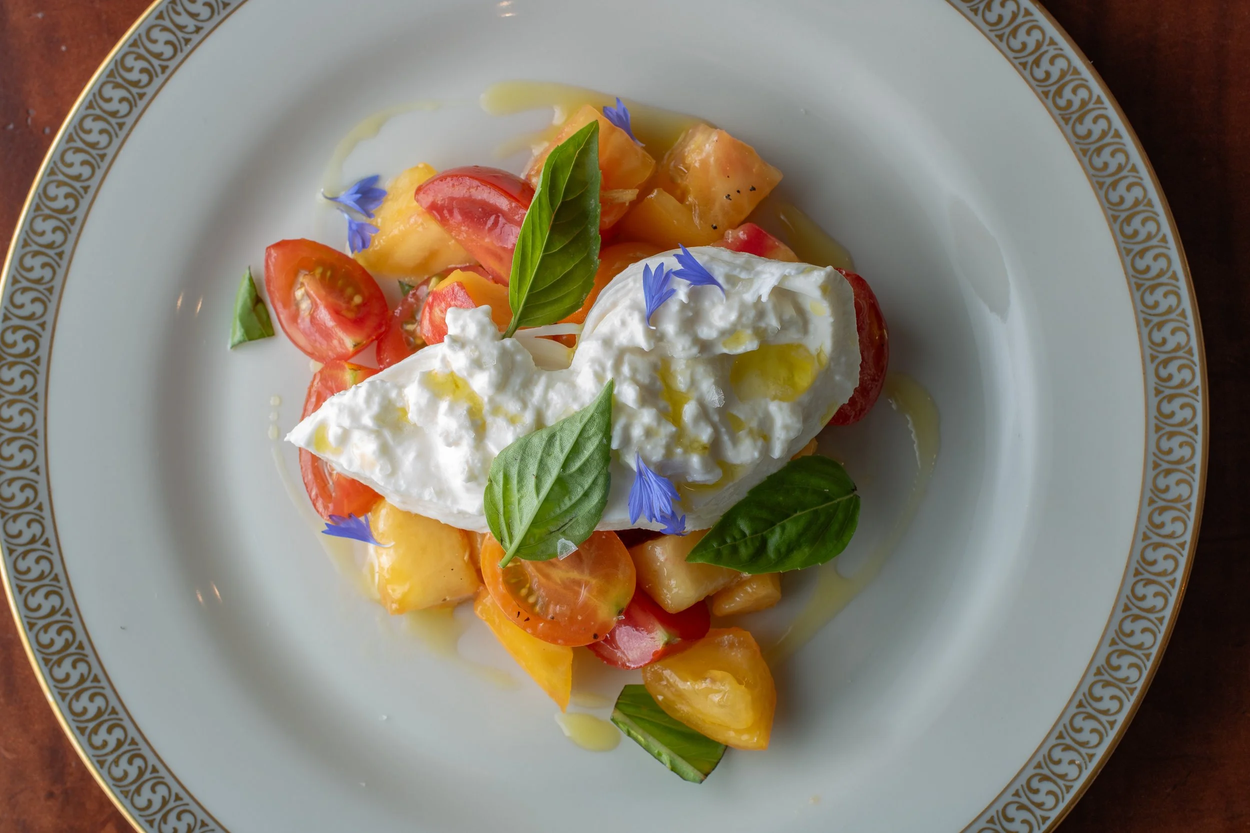 A plate of fresh tomato salad topped with dollops of mozzarella cheese, garnished with basil leaves and edible blue flower petals, drizzled with olive oil, on a decorative white plate with gold trim.
