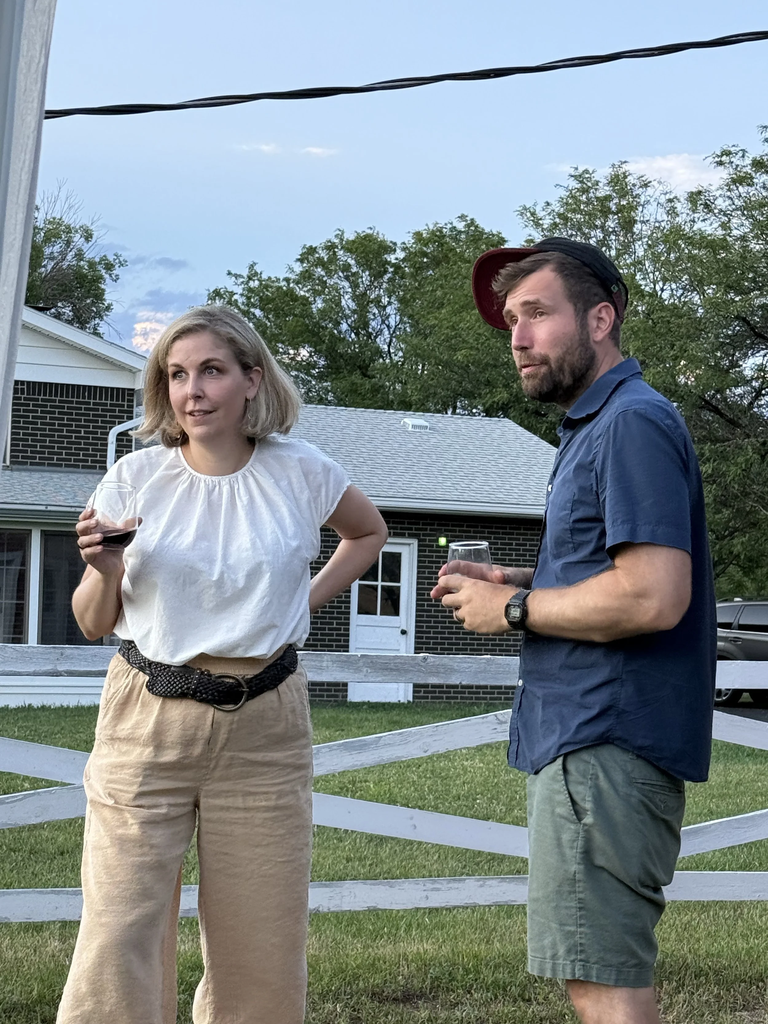 A woman with blonde hair wearing a white blouse and beige pants holding a glass of red wine, talking to a man with beard wearing a blue shirt, green shorts, and a red cap turned backward, holding a glass of wine, outdoors in a yard with a white fence