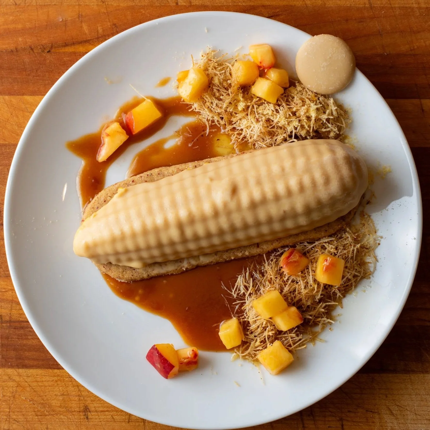 A white plate with a hot dog topped with cheese, surrounded by pineapple chunks, shredded meat, and a pool of brown sauce, on a wooden surface.