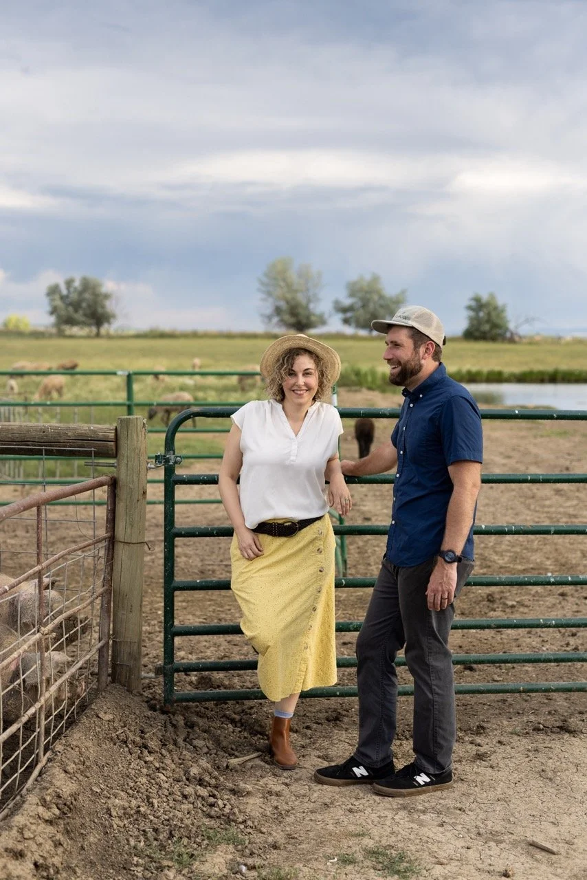 A woman in a white blouse, yellow skirt, and wide-brimmed hat leaning against a fence, smiling, next to a man in a blue shirt and gray cap, standing on a farm with a grassy field, trees, and a pond in the background.