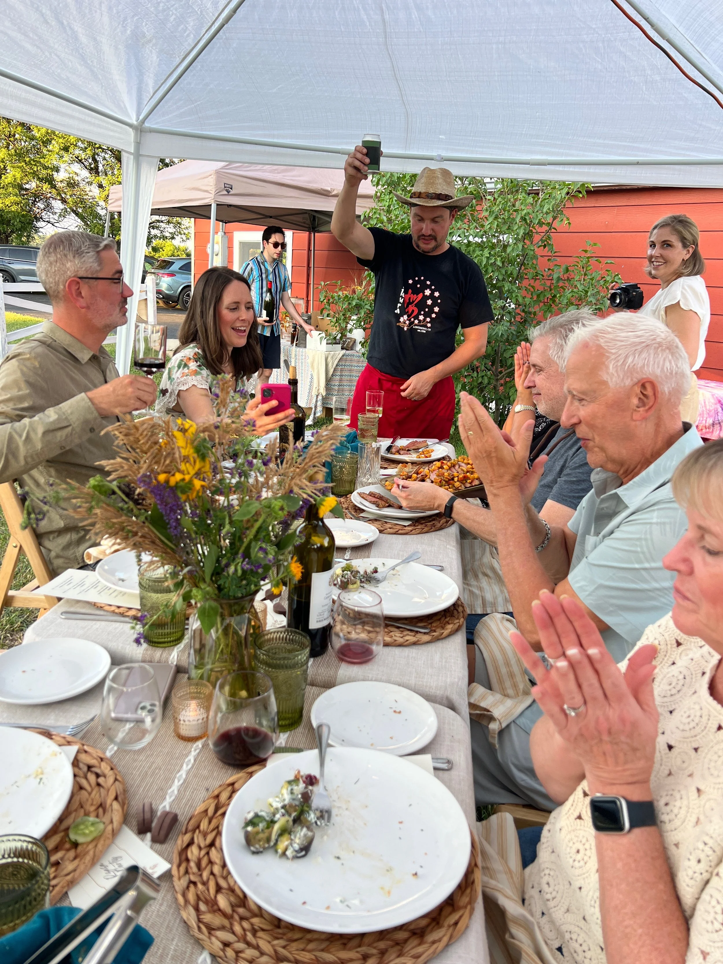 People gathered around a dining table under a canopy, celebrating outdoors with food and drinks, a man wearing a cowboy hat standing and taking a photo, and others smiling and clapping.