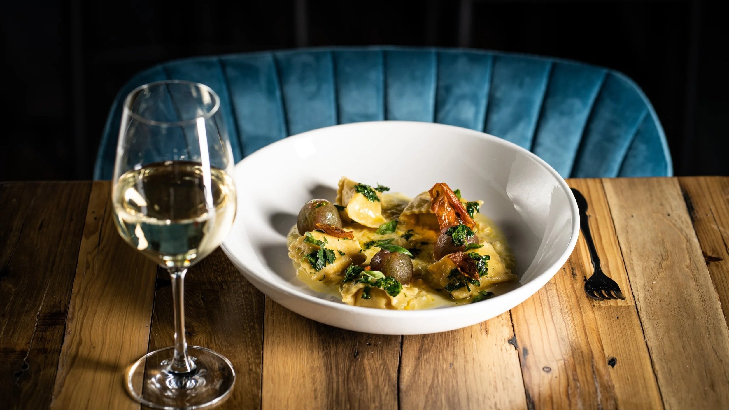 A bowl of pasta with greens and mushrooms on a wooden table, accompanied by a glass of white wine, with a blue velvet chair in the background.