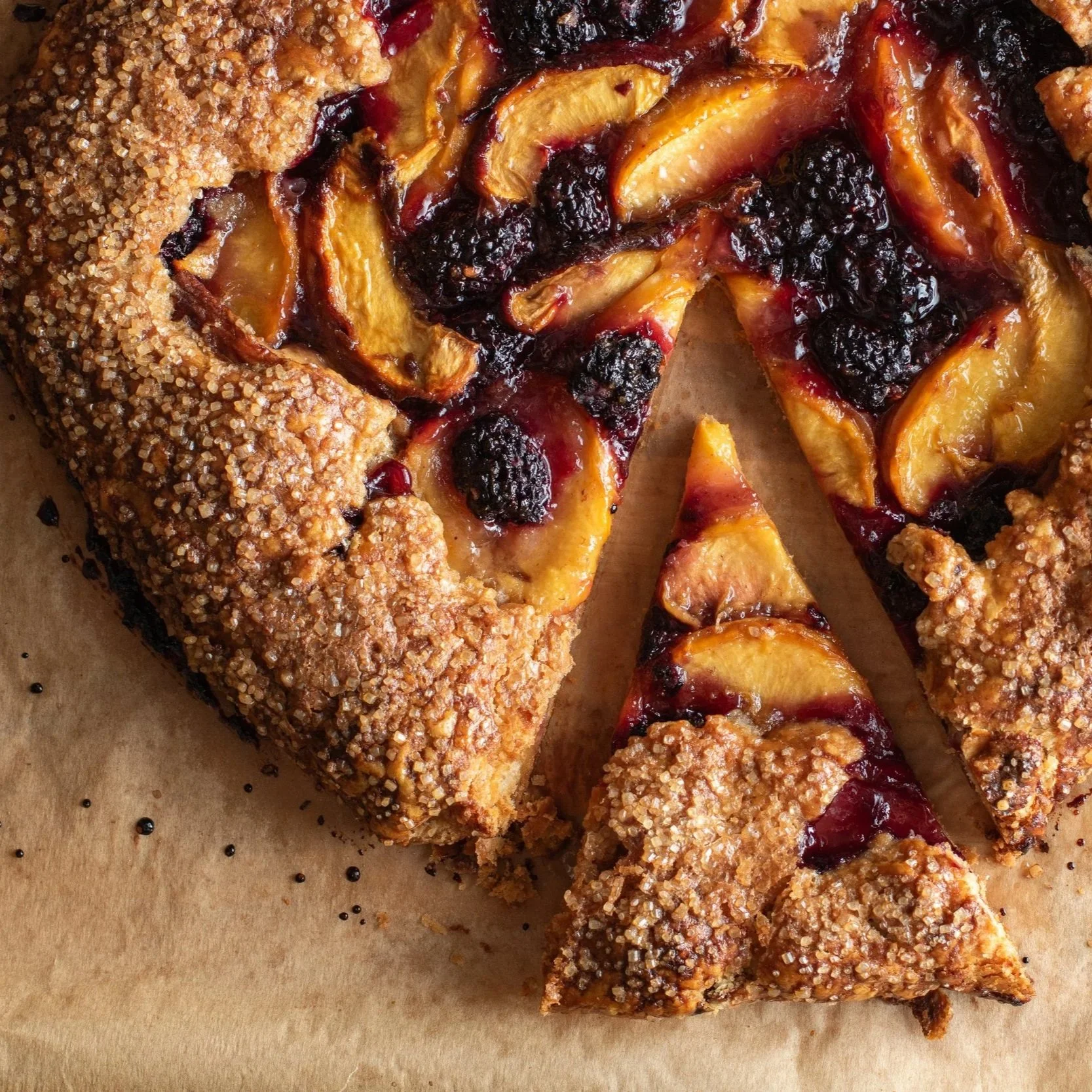 A sliced fruit tart with a crunchy sugar-coated crust, filled with peach slices and topped with a mix of blackberries and a fruit glaze.