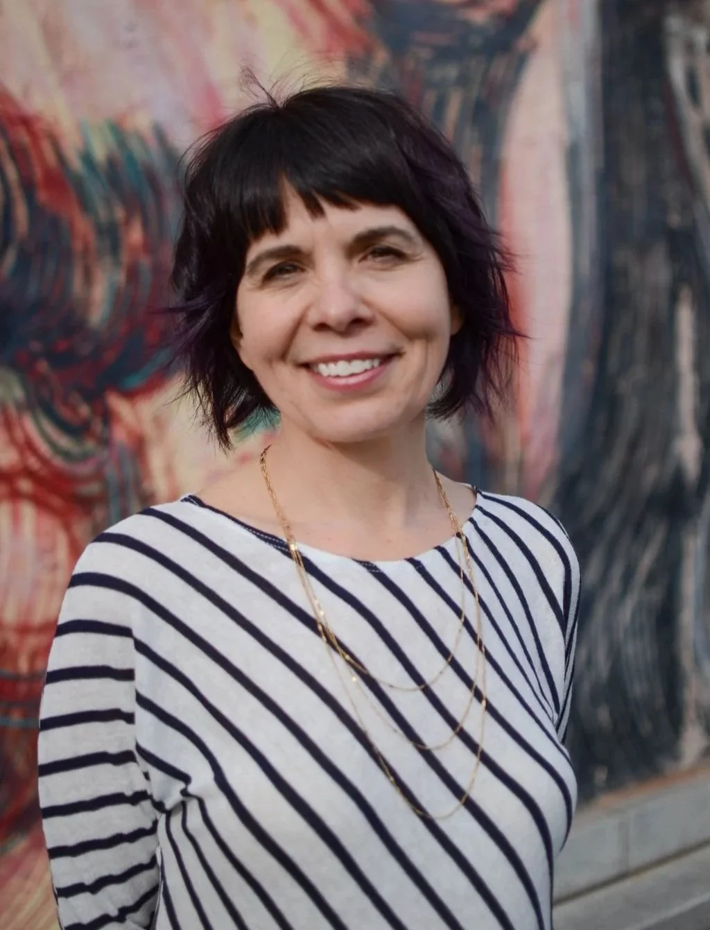 A woman with short dark hair and a striped shirt smiling in front of a colorful abstract painting.