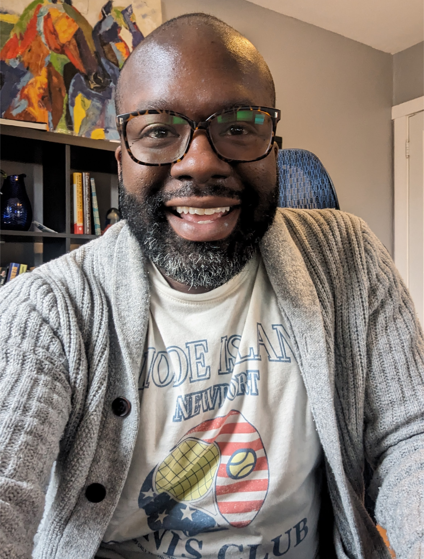 A smiling man with glasses, a beard, and a mustache, wearing a light gray cardigan over a graphic T-shirt, sitting in a room with a bookshelf and colorful artwork in the background.